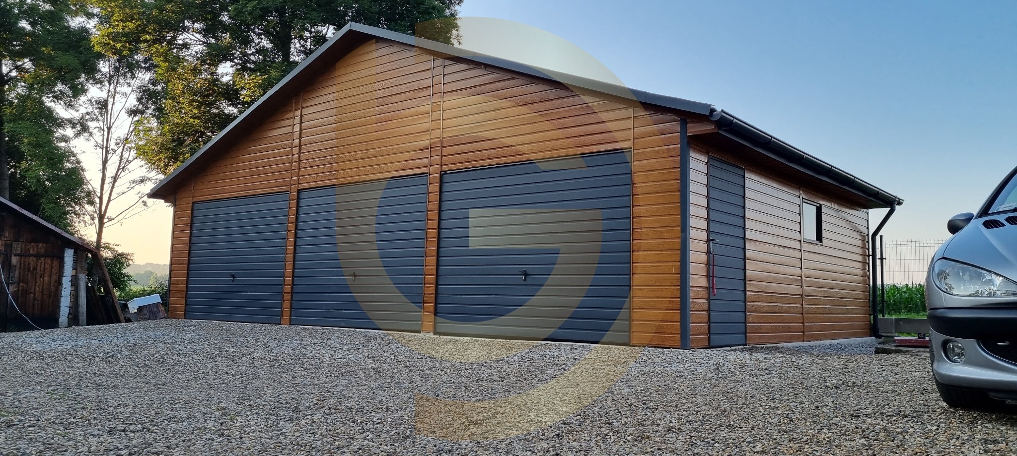 Drewniany garaż z trzema szarymi bramami segmentowymi, widok z zewnątrz na żwirowym podłożu, fragment srebrnego samochodu w kadrze.