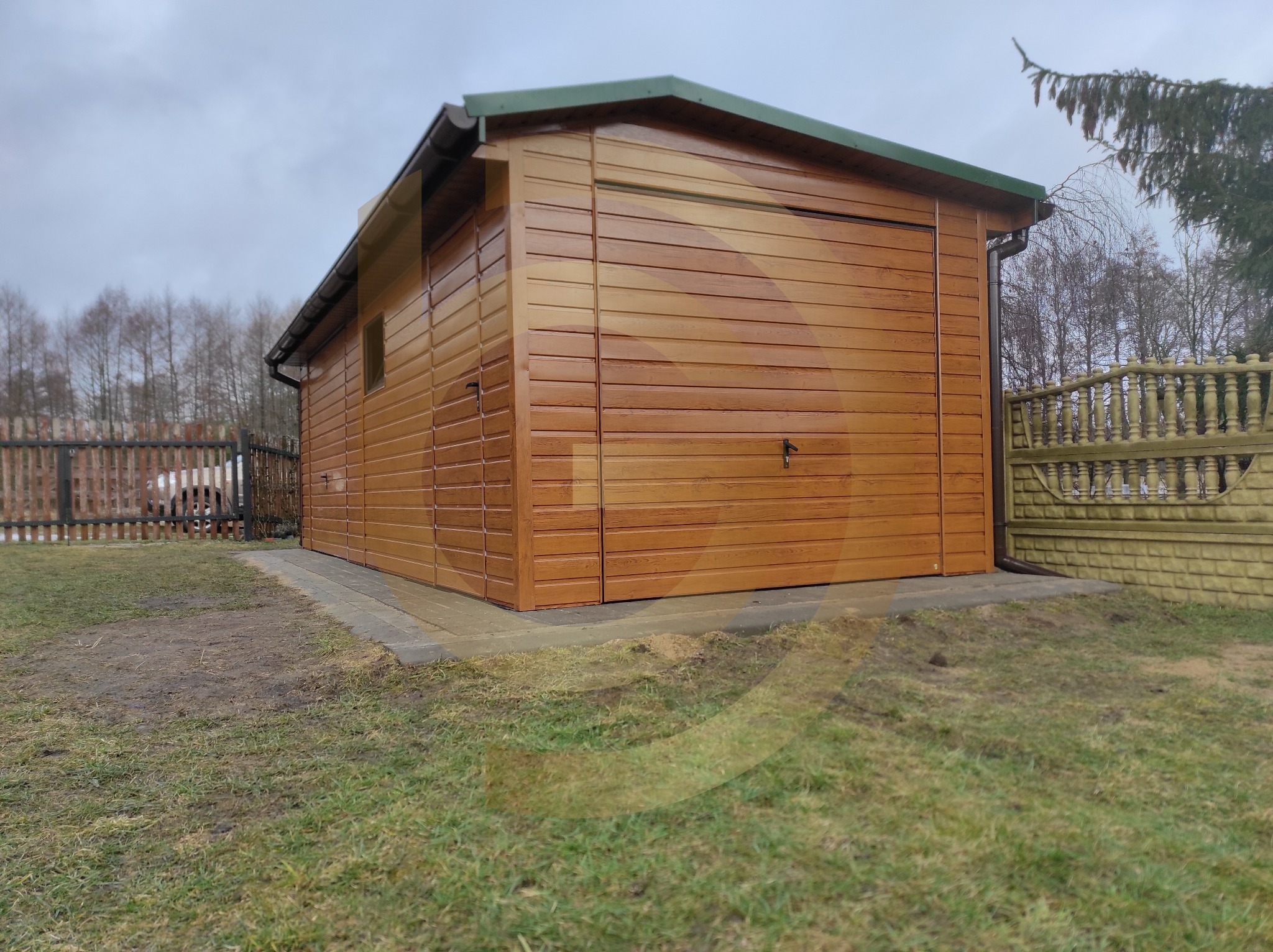 Drewniany garaż blaszany z segmentową bramą garażową, brązowe panele poziome, zielony dach, widoczny fragment trawnika i ogrodzenia.