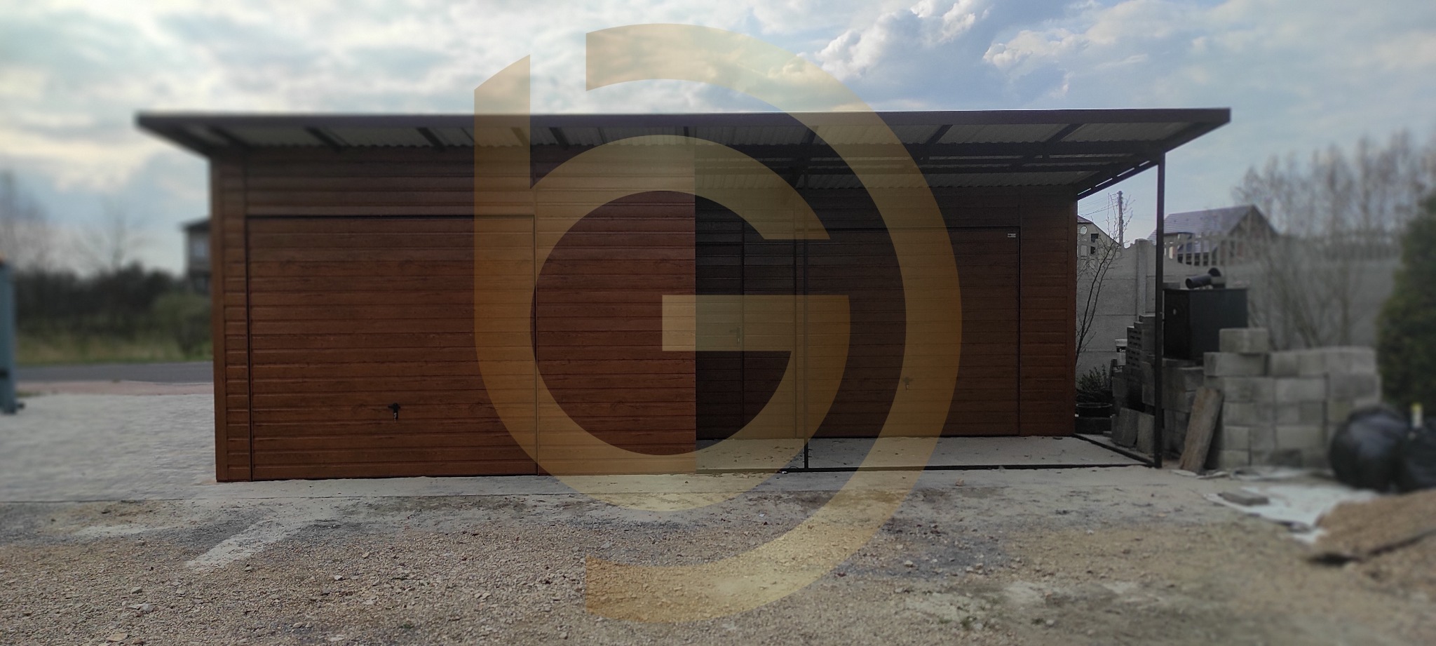 Brązowy garaż z dwoma bramami rolowanymi, blaszany dach, widok z zewnątrz na podwórku z kostką brukową i stertą betonowych bloków w tle.