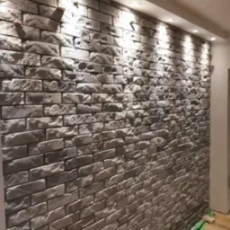 Ściana wewnętrzna wyłożona dekoracyjną cegłą w odcieniach szarości, oświetlona punktowymi lampami sufitowymi, z widocznym fragmentem korytarza w tle.