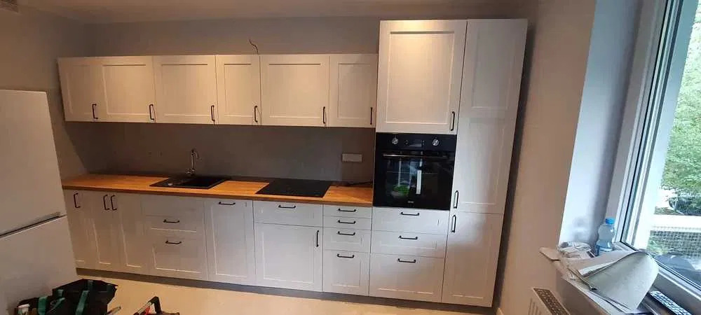 Jasna kuchnia z drewnianym blatem, czarnymi uchwytami i sprzętem AGD, widok z boku na okno z butelką wody mineralnej na parapecie.