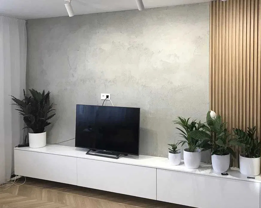 Szara ściana z efektem betonu, drewniane lamele na ścianie, biała szafka RTV z roślinami w doniczkach, telewizor i parkiet w jodełkę w nowoczesnym salonie.