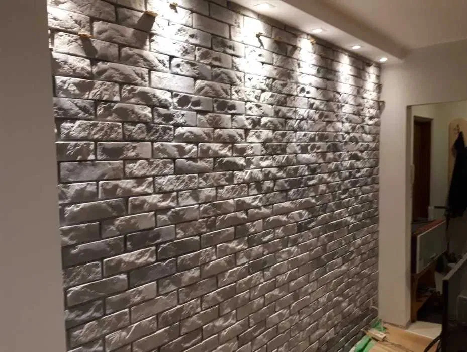 Ściana wewnętrzna wyłożona dekoracyjną cegłą w odcieniach szarości, oświetlona punktowymi lampami sufitowymi, z widocznym fragmentem korytarza w tle.