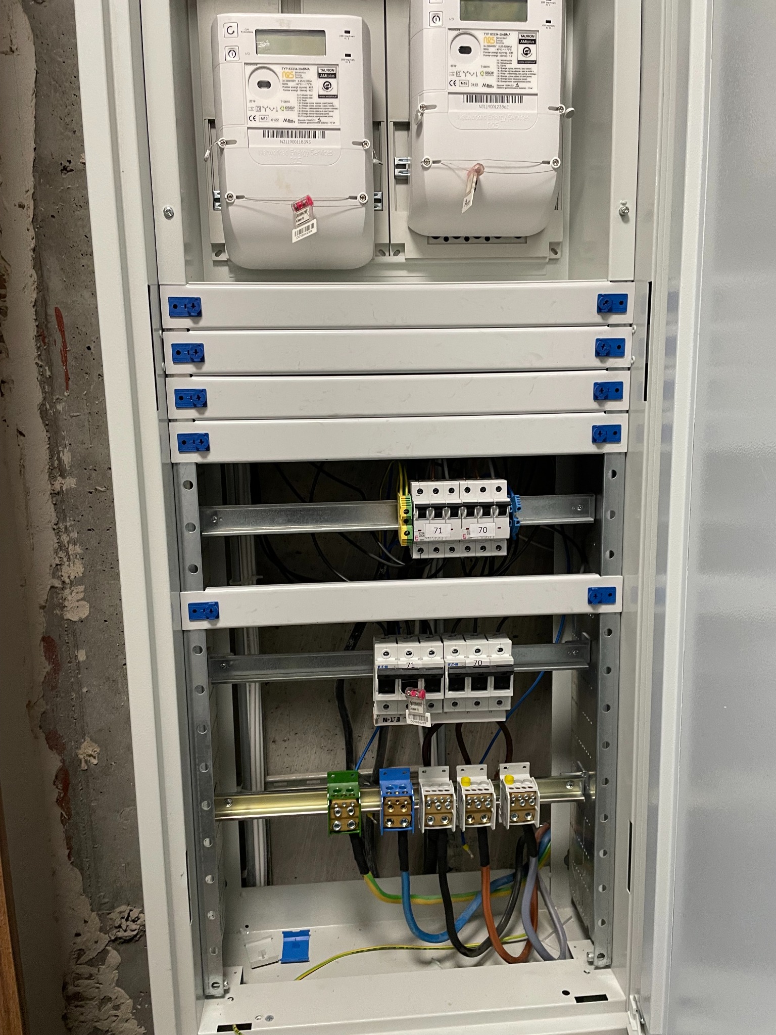 Otwarta szafa elektryczna z dwoma licznikami energii, bezpiecznikami i okablowaniem, widoczne złącza i listwy zaciskowe.