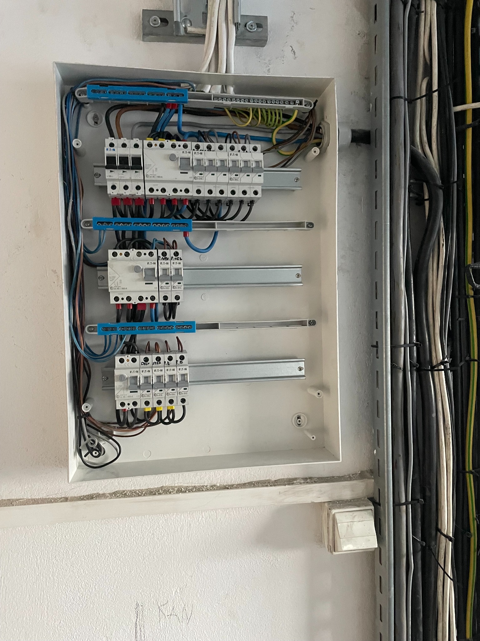 Otwarta skrzynka elektryczna z zamontowanymi bezpiecznikami i przewodami. Widoczne kable w ścianie. Biała ściana z szarą listwą przypodłogową.