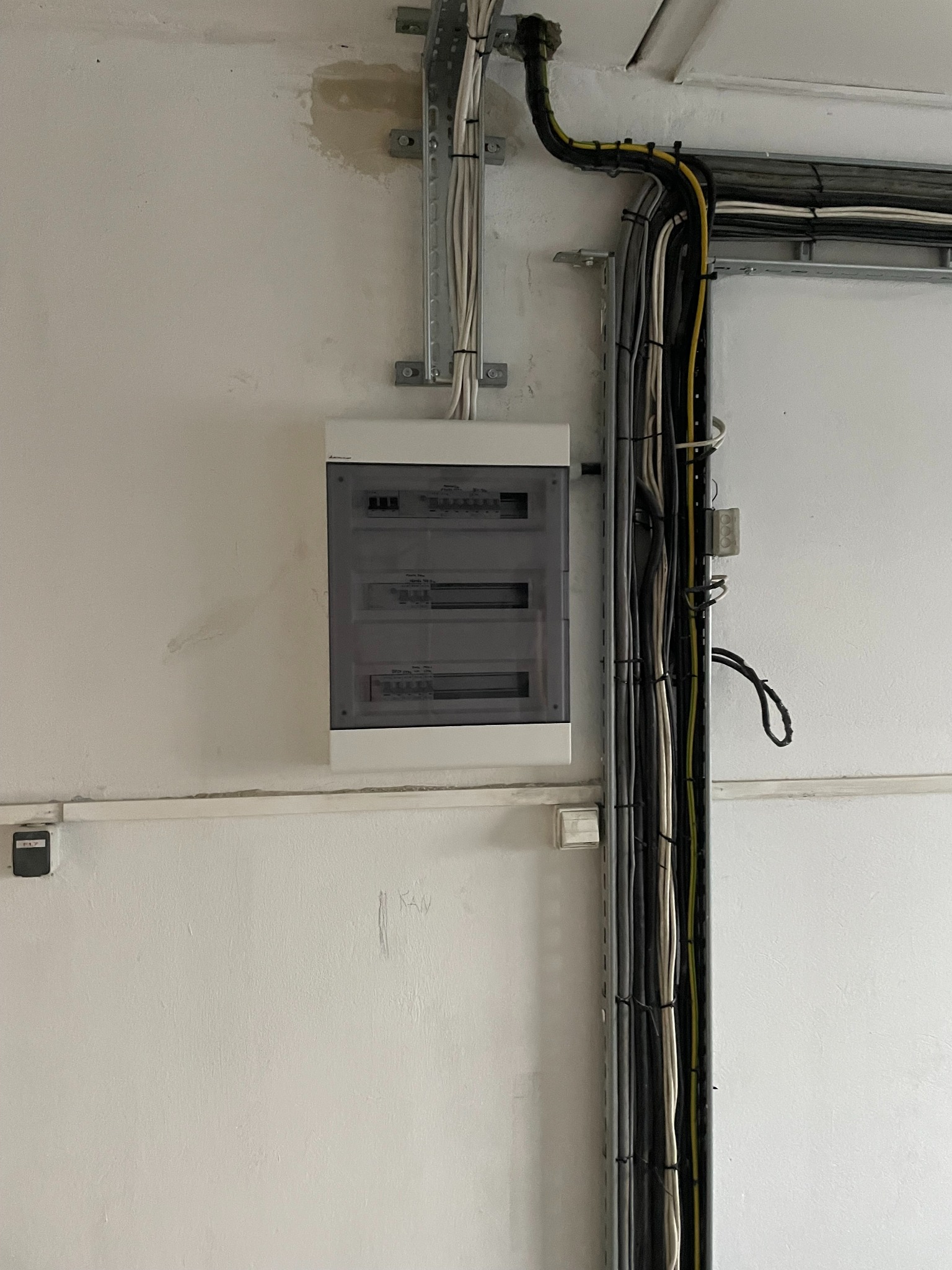 Szara skrzynka elektryczna z zainstalowanymi bezpiecznikami na białej ścianie. Nad skrzynką widoczne kable w korytkach. Widoczne ślady po zalaniu.