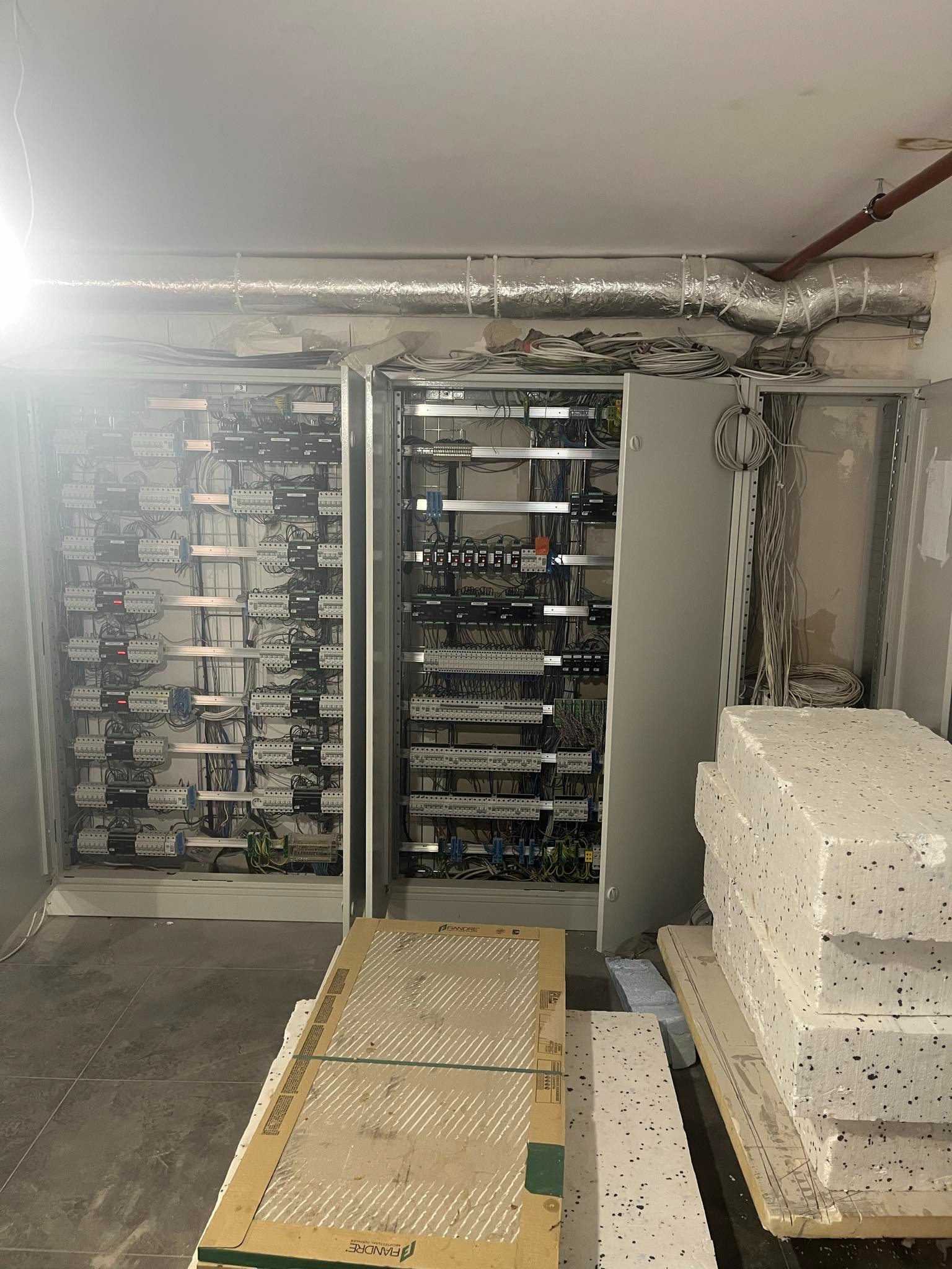 Rozdzielnice elektryczne w szarej obudowie, widoczne liczne kable i podłączenia. Na podłodze płyty ceramiczne i styropian. Instalacja w trakcie realizacji.