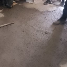 Grzegorz sitek - Świeżo wylana, mokra posadzka przemysłowa w trakcie poziomowania, widoczne fragmenty butów robotników i ściana z białymi płytkami.