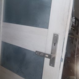 Grzegorz sitek - Białe drzwi wewnętrzne z poziomymi szarymi szybami i metalową klamką, widoczne ślady montażu w tle.