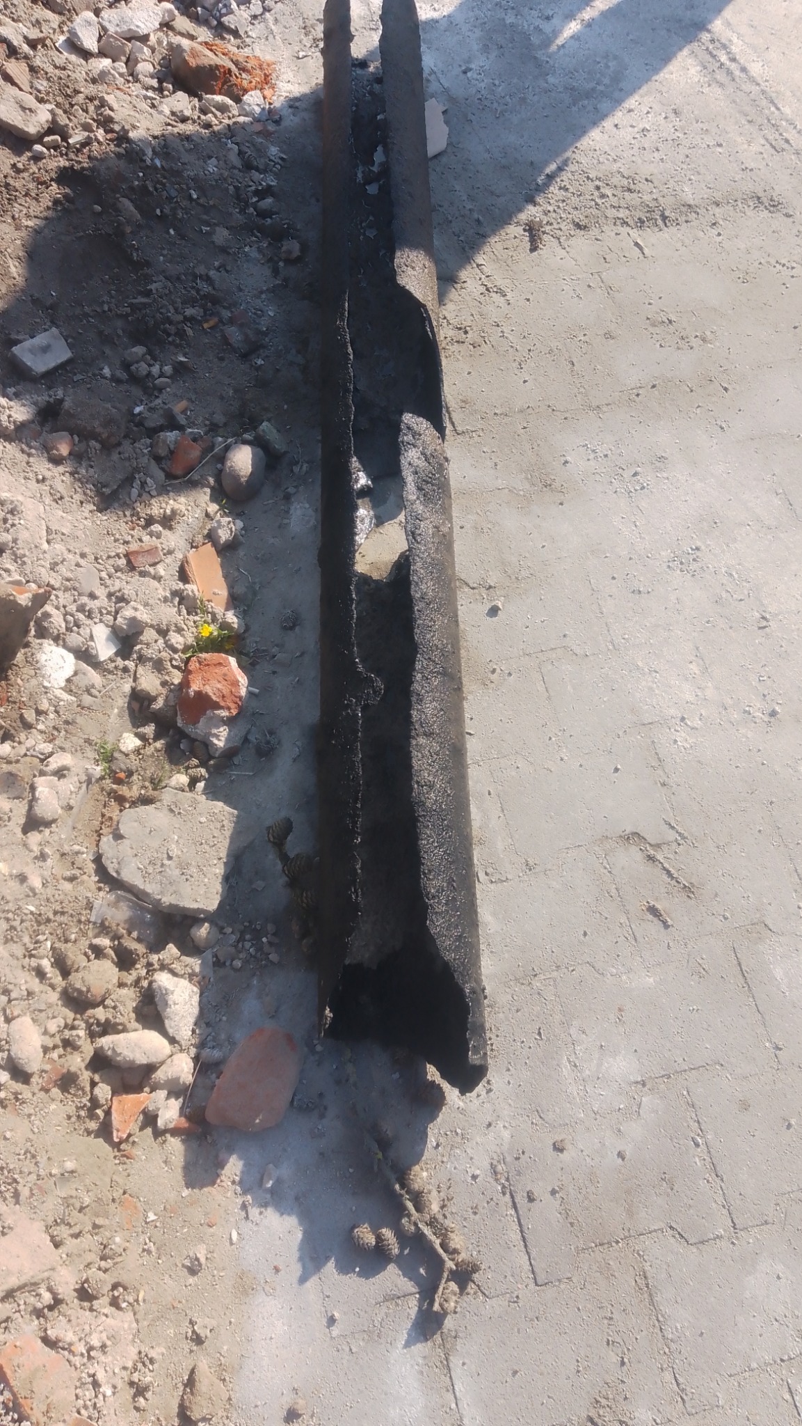 Uszkodzona, czarna rura leżąca na betonowej powierzchni z gruzem i kawałkami cegieł w pobliżu.