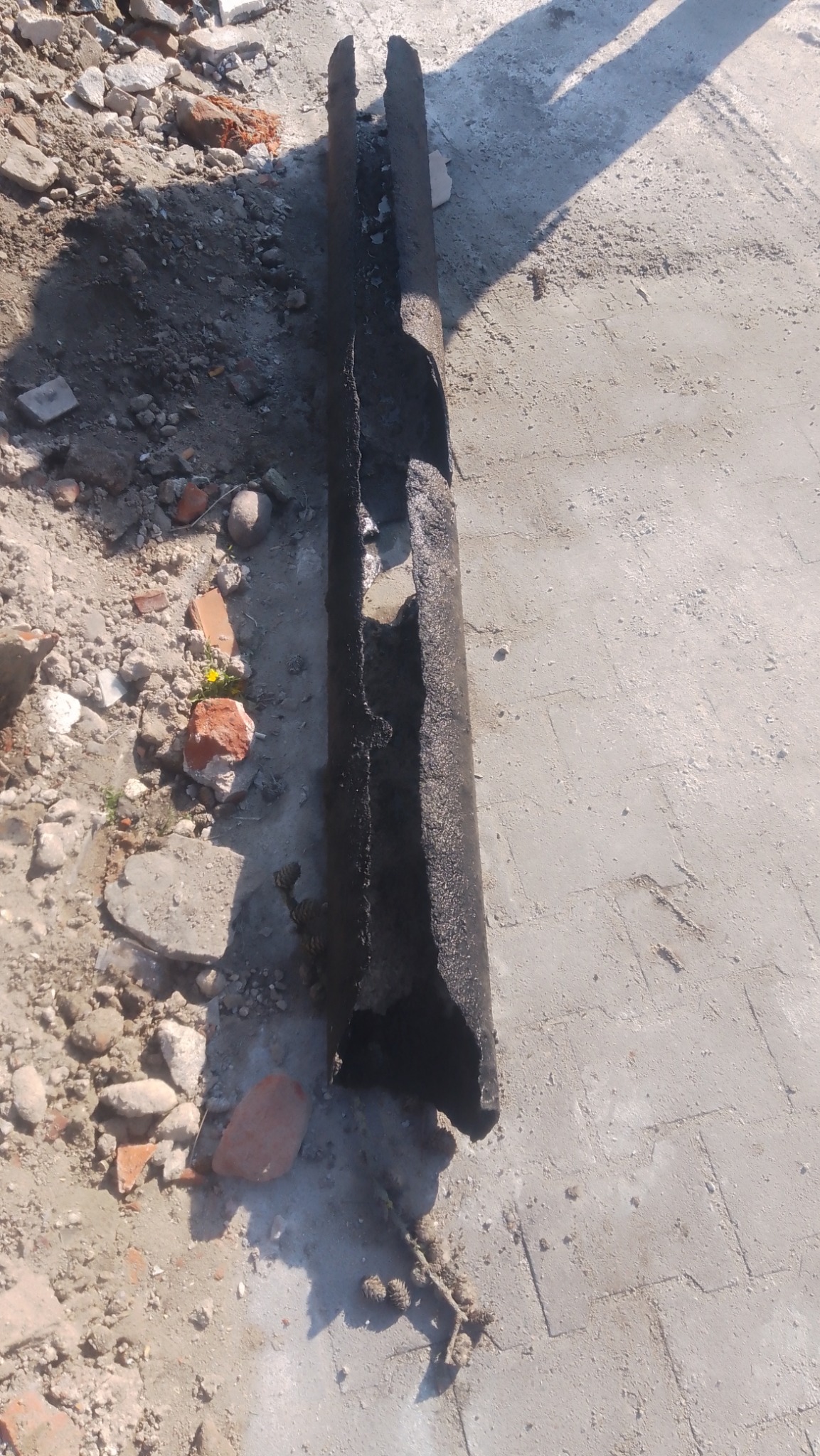 Uszkodzony fragment rury z czarną izolacją leżący na betonowej powierzchni wśród gruzu i kamieni.