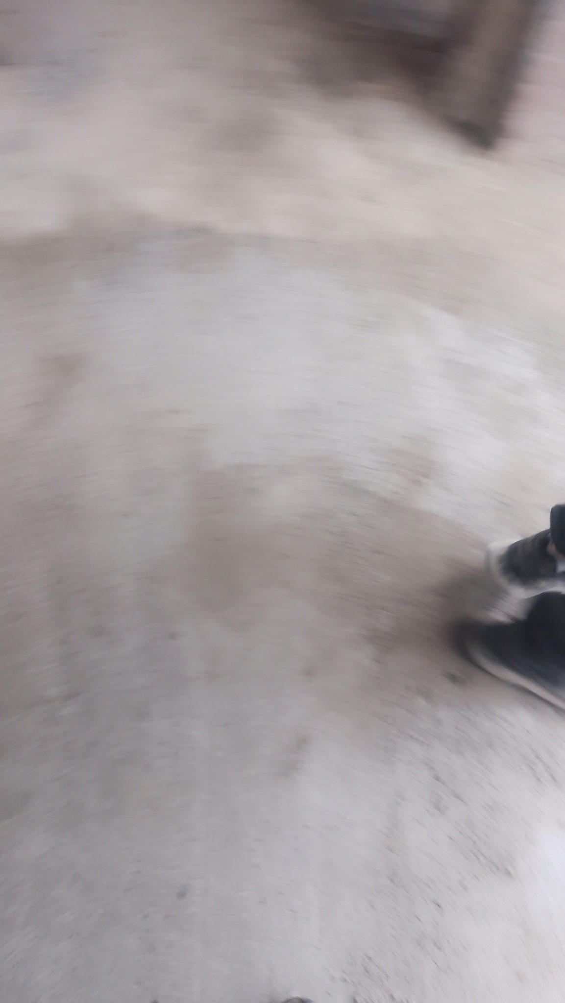 Rozmazane ujęcie szarej, prawdopodobnie betonowej posadzki, z fragmentem buta w prawym dolnym rogu.