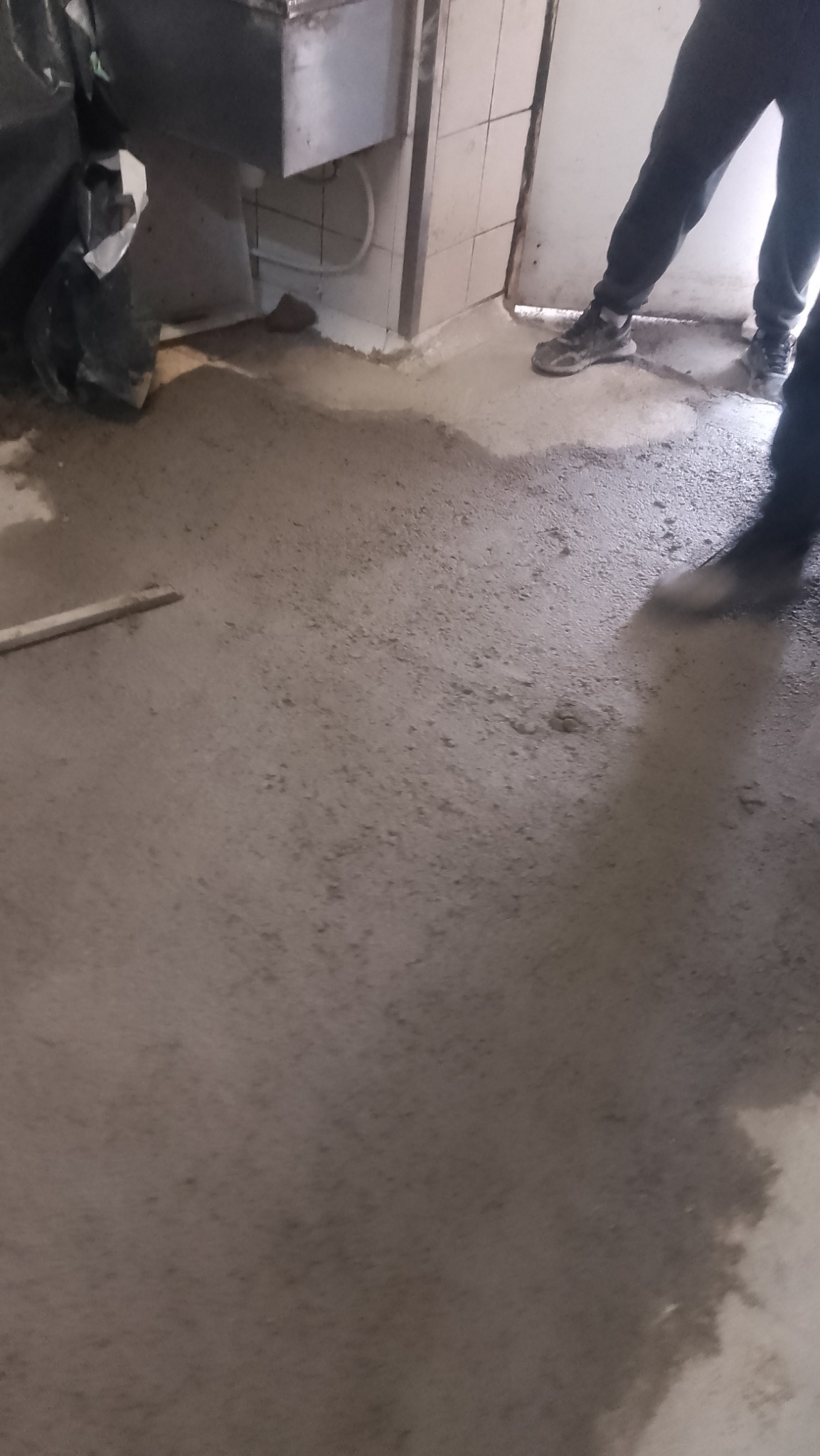 Świeżo wylana, mokra posadzka przemysłowa w trakcie poziomowania, widoczne fragmenty butów robotników i ściana z białymi płytkami.