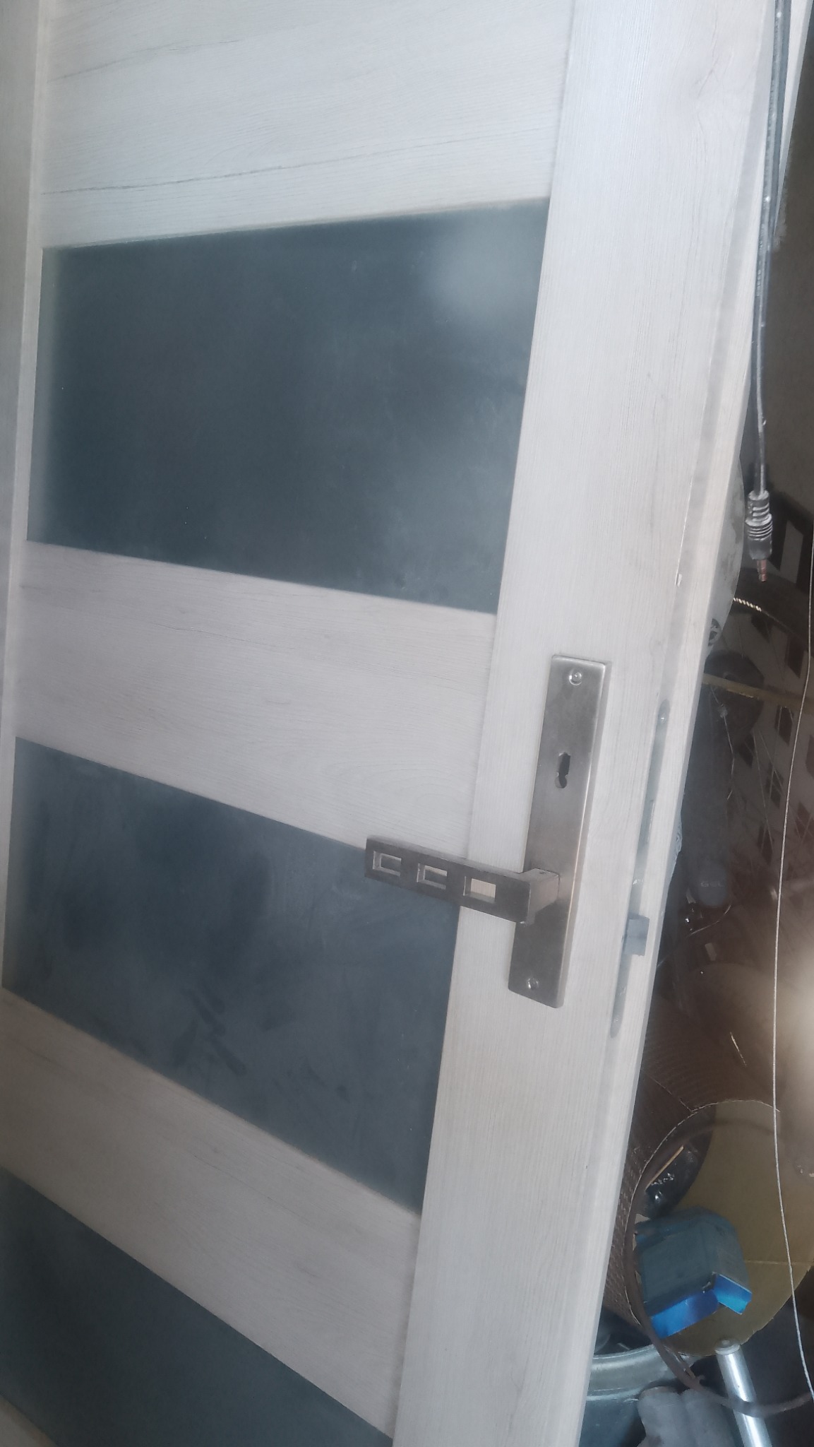 Białe drzwi wewnętrzne z poziomymi szarymi szybami i metalową klamką, widoczne ślady montażu w tle.