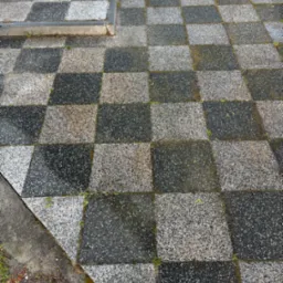 Szachownica z kostki brukowej w odcieniach szarości, miejscami wilgotna i porośnięta mchem, widoczne fragmenty trawy i ziemi na obrzeżach.