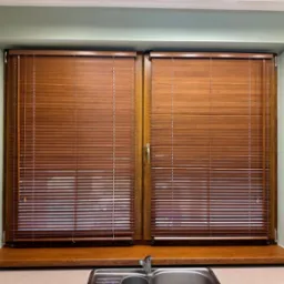 Okna kuchenne z drewnianymi żaluzjami w kolorze orzecha, zamontowane nad zlewem z baterią, widoczne beżowe płytki i gniazdko elektryczne.