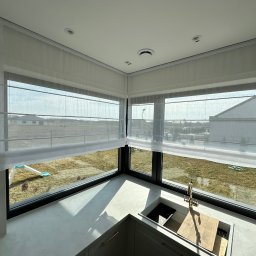 Krzysztof Kobiela TWÓJ STYL - Jasna kuchnia z narożnym oknem przesłoniętym białymi, półprzezroczystymi roletami rzymskimi, widok na ogród z huśtawką.