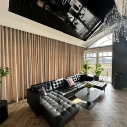 Beżowe zasłony w salonie z czarną skórzaną sofą, roślinami doniczkowymi i parkietem ułożonym w jodełkę, widok z perspektywy podłogi.