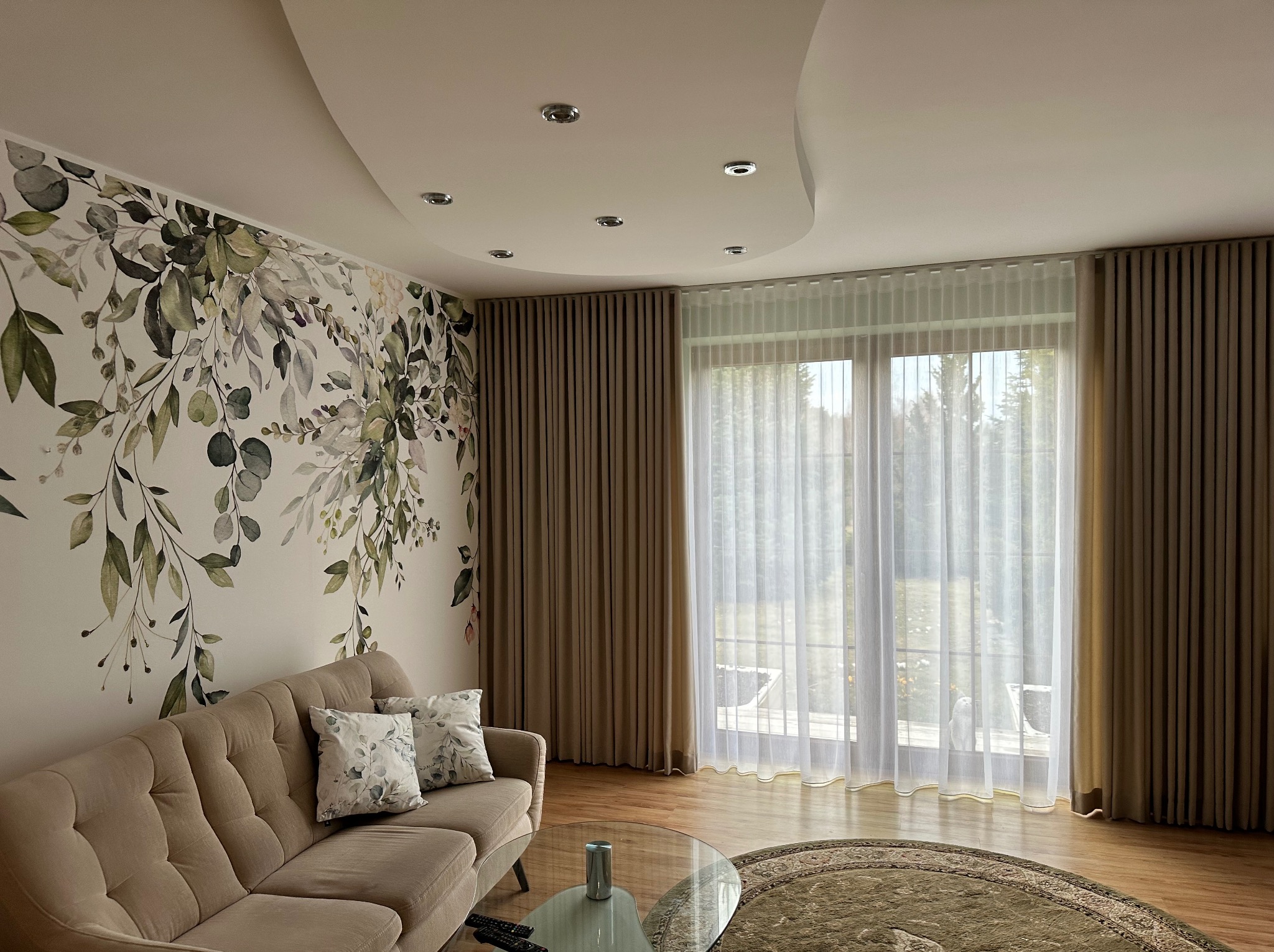 Eleganckie wnętrze salonu z beżową sofą, dekoracyjną tapetą w motywy roślinne i zasłonami w odcieniach beżu, wpadające przez okno światło dzienne.