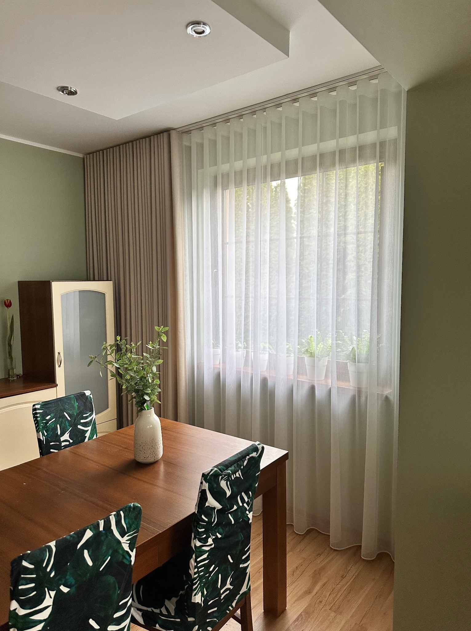 Jadalnia z drewnianym stołem, krzesłami w zielone liście i oknem z długimi, białymi, zwiewnymi zasłonami, za którymi widać zieleń drzew. W tle beżowe zasłony i komoda.