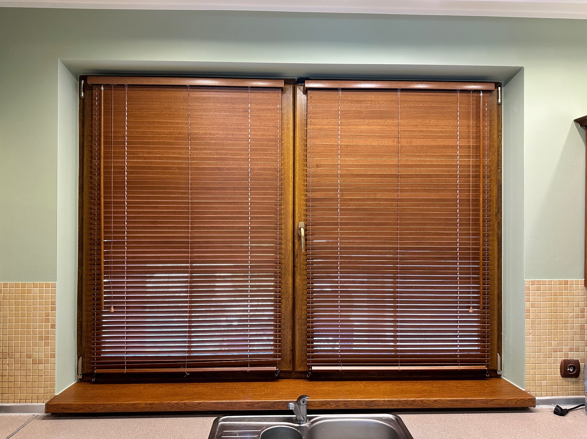 Okna kuchenne z drewnianymi żaluzjami w kolorze orzecha, zamontowane nad zlewem z baterią, widoczne beżowe płytki i gniazdko elektryczne.