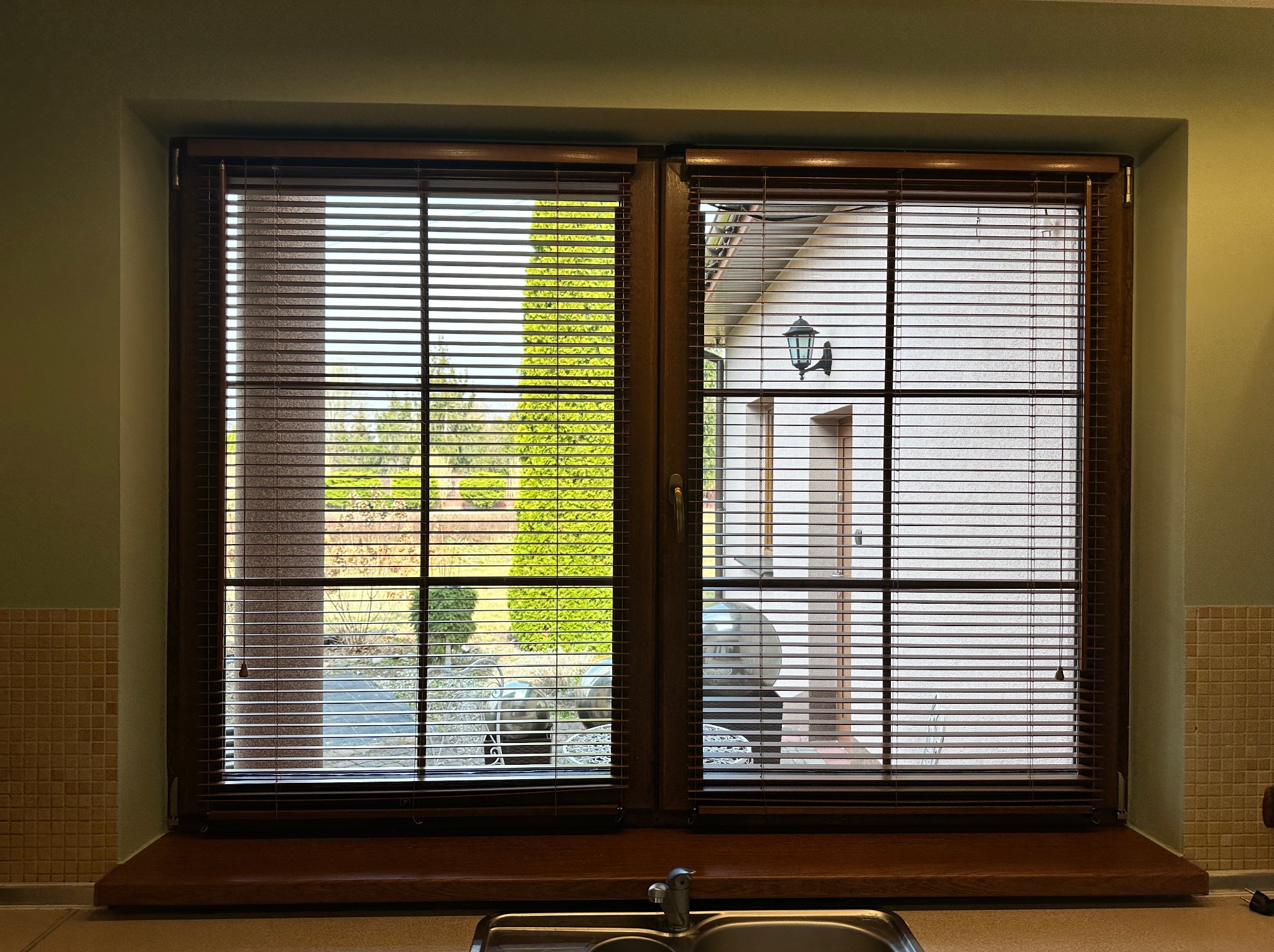 Okna z brązowymi żaluzjami poziomymi, widok na ogród i dom z lampą ścienną.