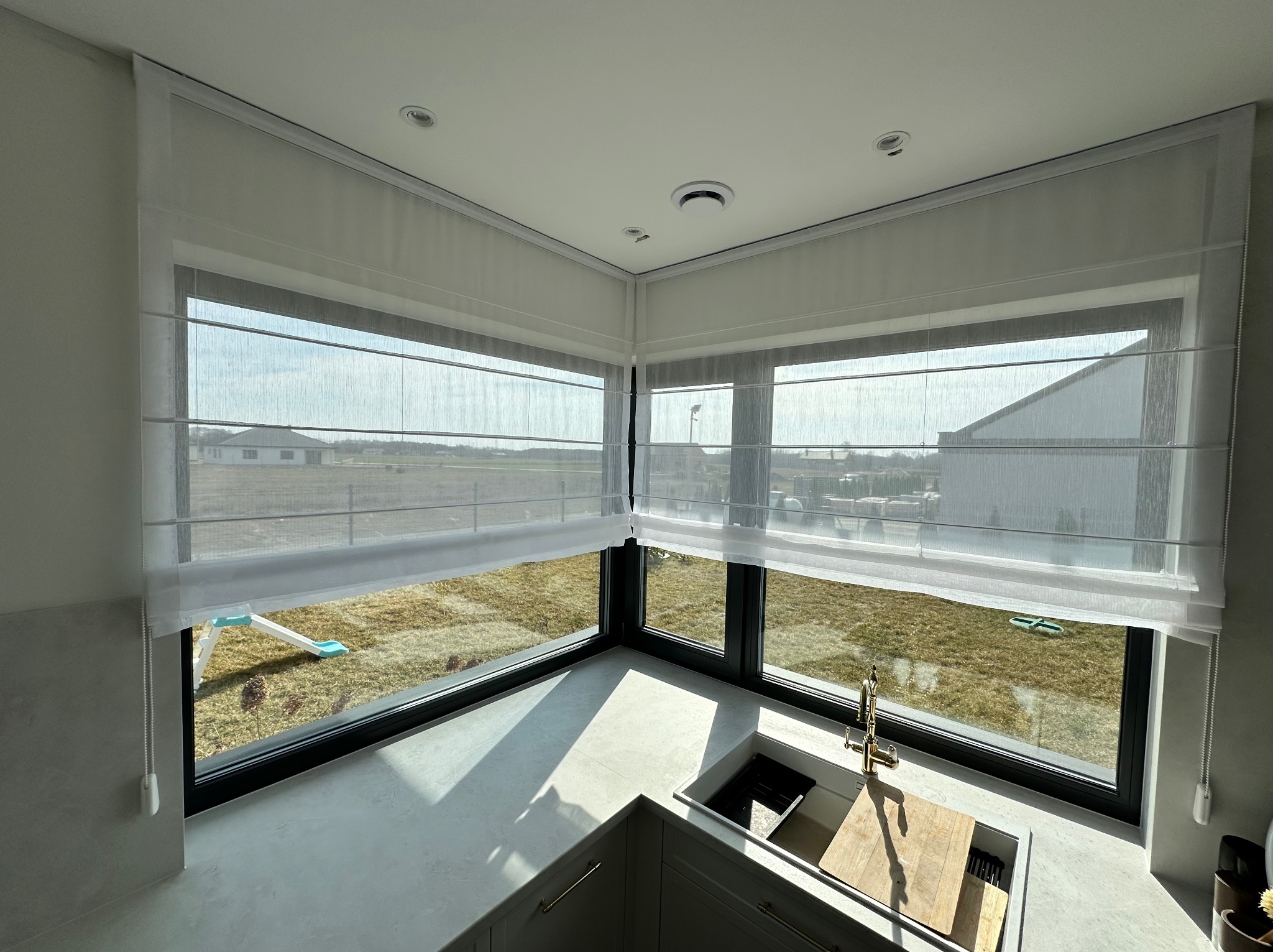 Jasna kuchnia z narożnym oknem przesłoniętym białymi, półprzezroczystymi roletami rzymskimi, widok na ogród z huśtawką.