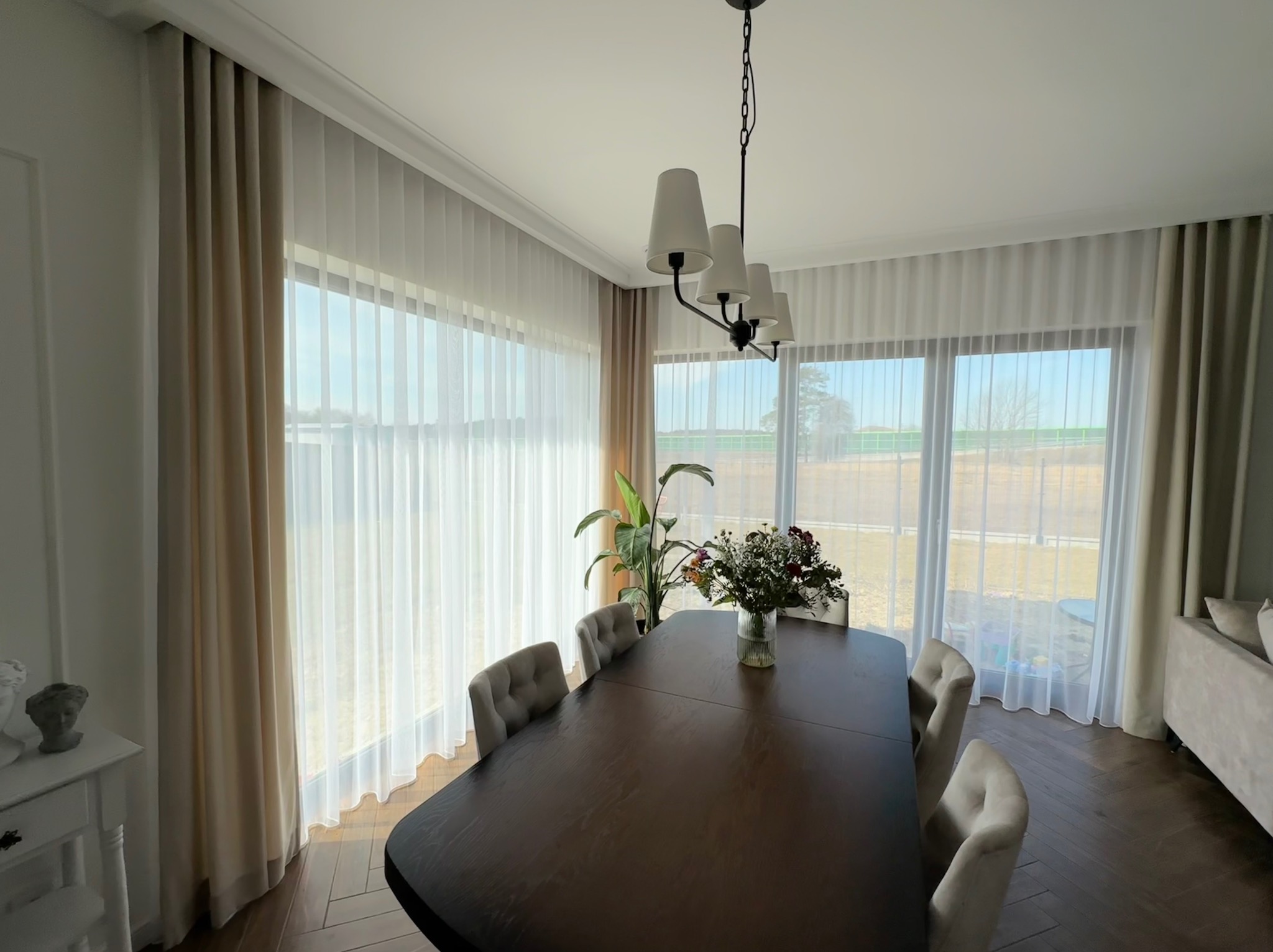 Jasne, przestronne wnętrze jadalni z dużym drewnianym stołem, szarymi krzesłami i zwiewnymi, beżowymi zasłonami w oknach.