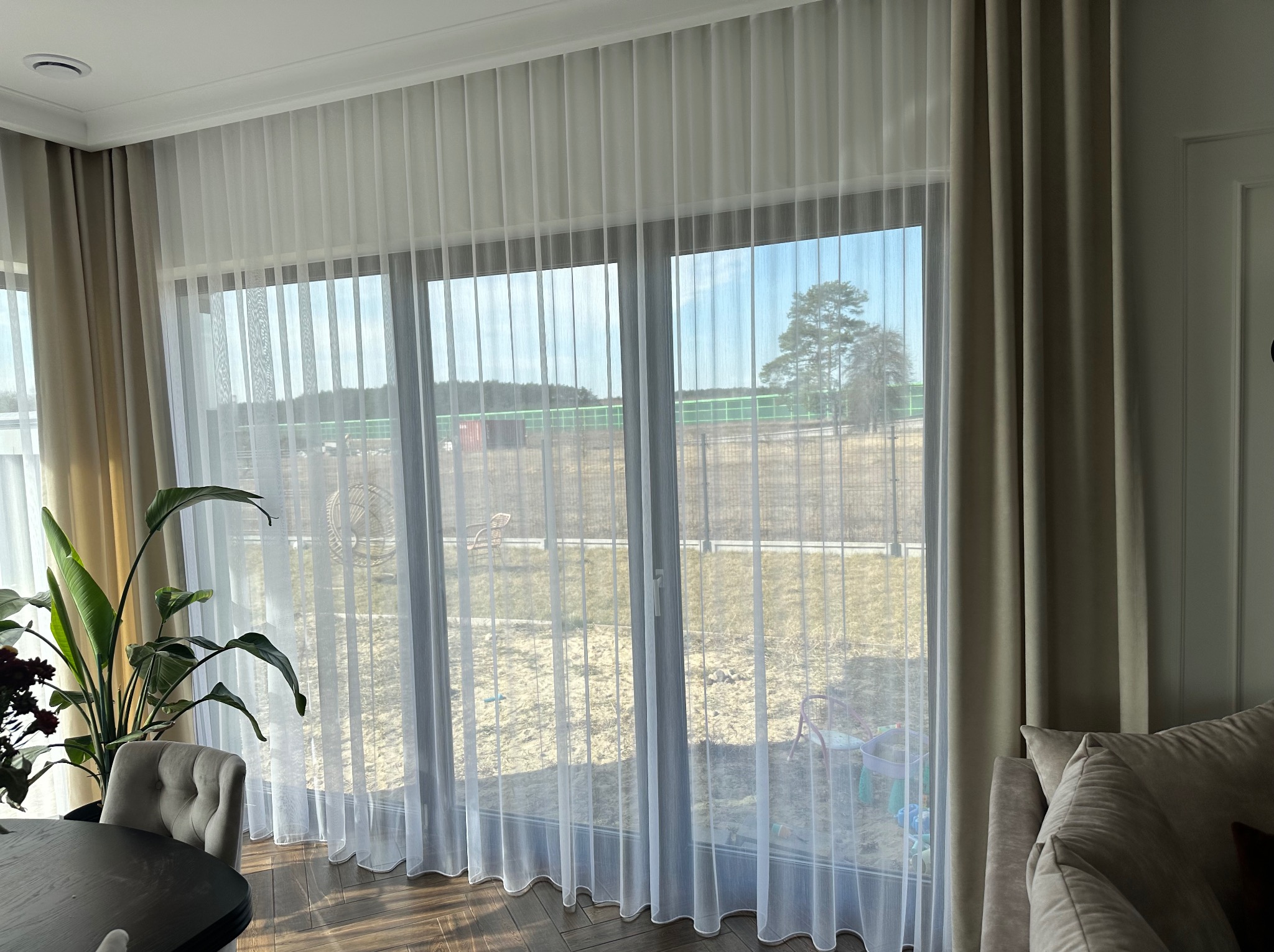 Delikatne, białe, marszczone firany w oknie, zestawione z beżowymi zasłonami po bokach, widok na zewnątrz z placem zabaw, fragment stołu z krzesłem i rośliną doniczkową w pomieszczeniu.