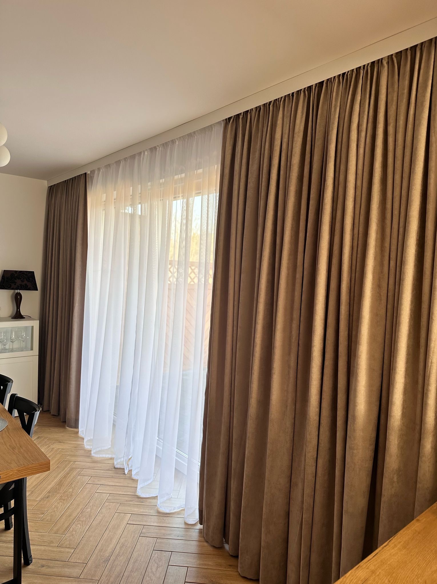 Beżowe, grube zasłony i białe, prześwitujące firany zawieszone na szynie sufitowej w jasnym salonie z parkietem w jodełkę.