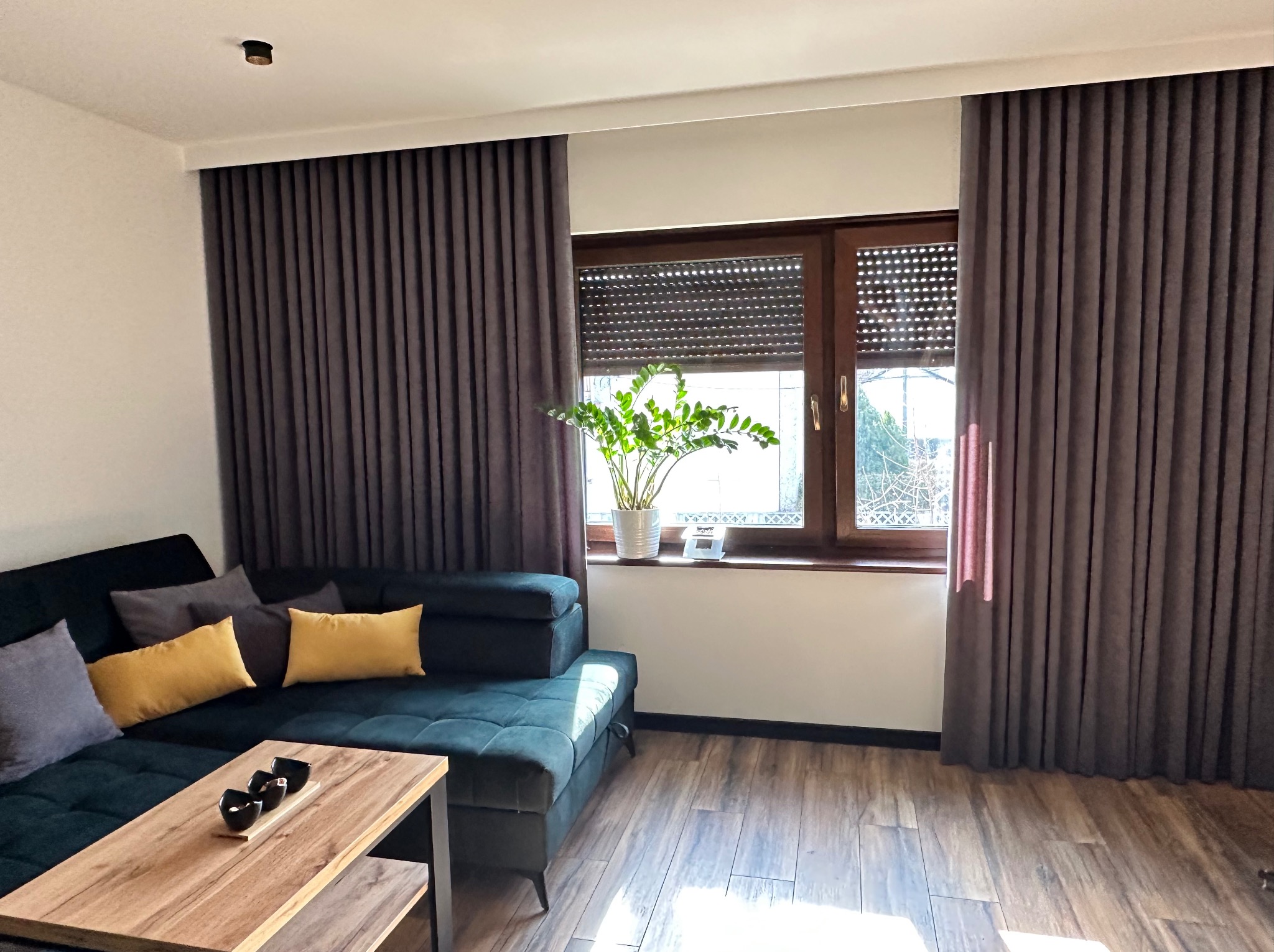 Nowoczesny salon z szarobrązowymi zasłonami, roletami zewnętrznymi, zieloną sofą i drewnianym stolikiem kawowym z czarnymi miseczkami.