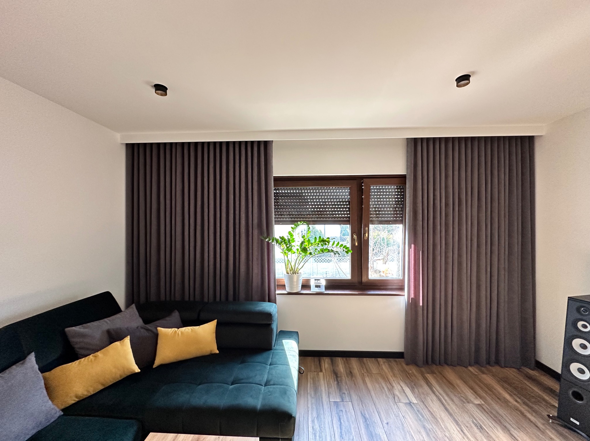 Zasłony w odcieniach szarości, z wyraźnymi pionowymi fałdami, zamontowane po bokach okna z drewnianą ramą i roletą, widoczne w salonie z ciemnozieloną sofą i drewnianą podłogą