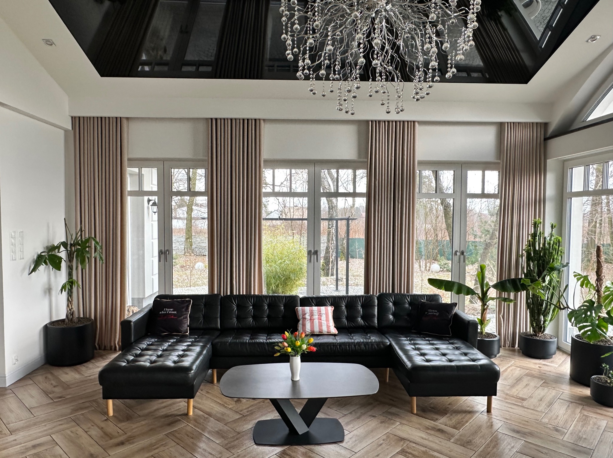 Elegancki salon z czarną skórzaną sofą modułową, beżowymi zasłonami, parkietem w jodełkę, roślinami doniczkowymi i nowoczesnym żyrandolem kryształowym