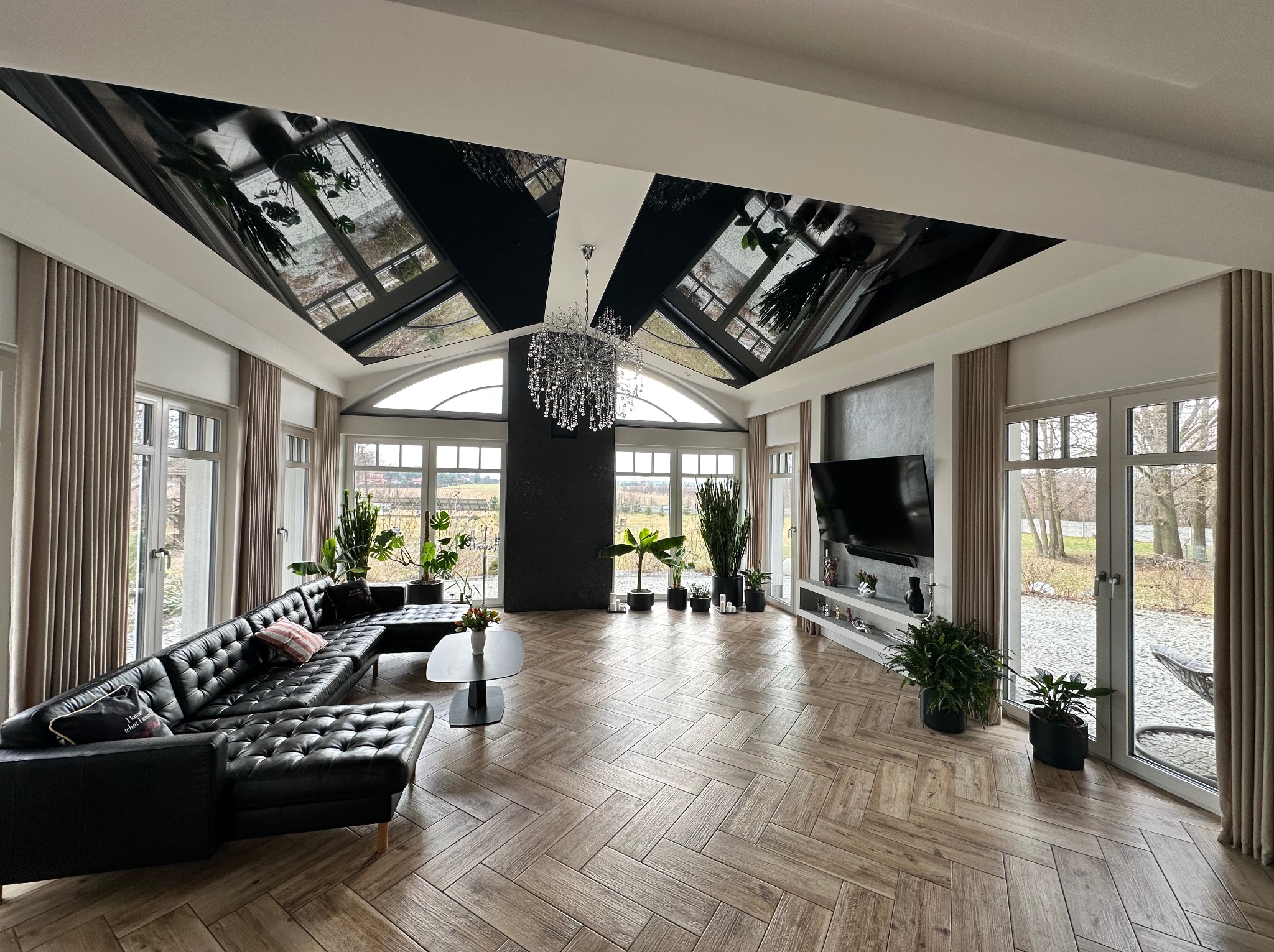 Przestronny salon z parkietem ułożonym w jodełkę, czarną skórzaną sofą narożną, wysokimi oknami z beżowymi zasłonami i lustrzanym sufitem z widocznym odbiciem roślin doniczkowych.