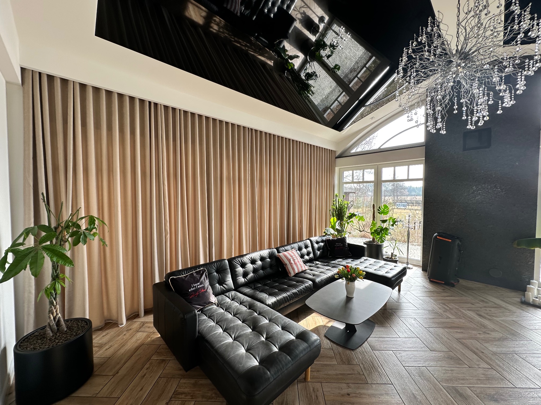 Beżowe zasłony w salonie z czarną skórzaną sofą, roślinami doniczkowymi i parkietem ułożonym w jodełkę, widok z perspektywy podłogi.