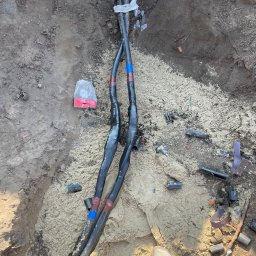 Elmarex Mariusz Kucharski - Dwa czarne kable elektryczne w wykopie, zabezpieczone taśmą izolacyjną, otoczone piaskiem i elementami montażowymi. Widoczny fragment buta osoby nadzorującej.