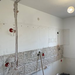 Elmarex Mariusz Kucharski - Białe przewody elektryczne w bruzdach na ścianie w trakcie montażu, widoczne puszki instalacyjne i fragment instalacji oświetleniowej pod sufitem.