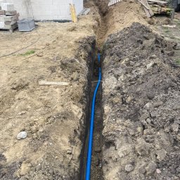 Elmarex Mariusz Kucharski - Niebieski karbowany przewód w wykopie ziemnym, prawdopodobnie do instalacji elektrycznej, na tle budowy domu z białego bloczka.