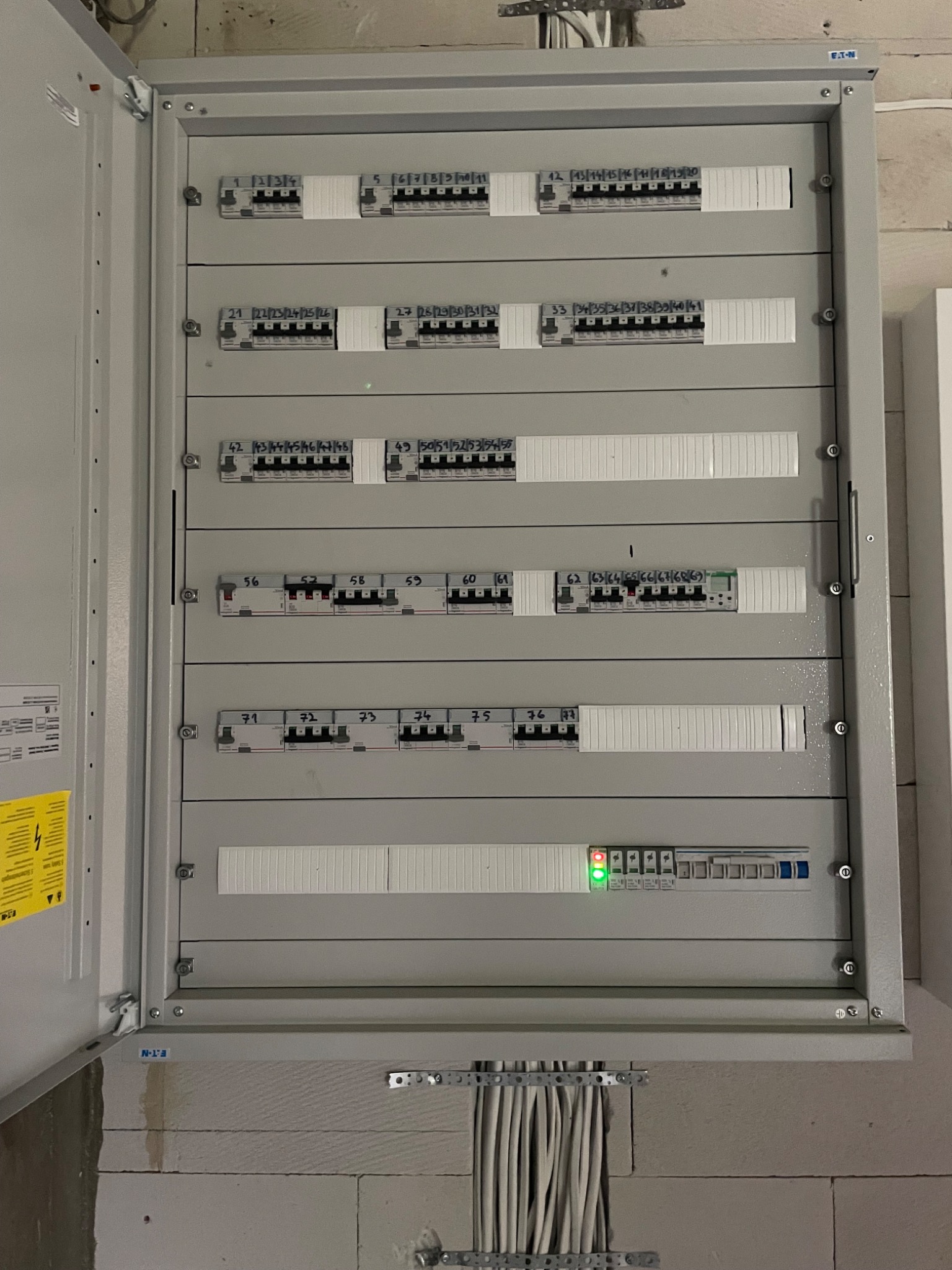 Otwarta szara skrzynka rozdzielcza z bezpiecznikami i numerowanymi wyłącznikami, zamontowana na ścianie z betonu komórkowego. Widoczne kable wchodzące do skrzynki.