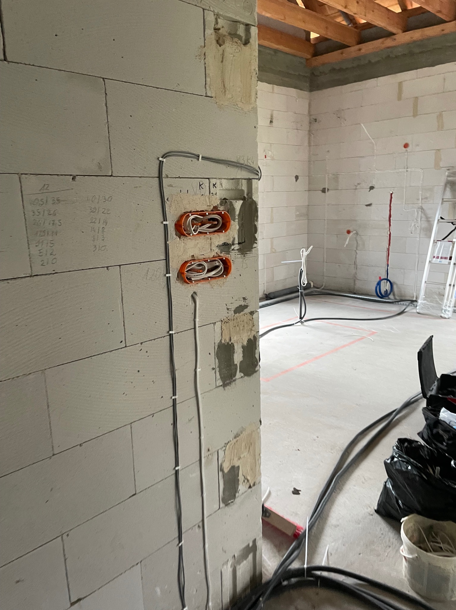 Dwa pomarańczowe gniazdka elektryczne w surowej ścianie z betonu komórkowego, z widocznymi przewodami. W tle instalacja elektryczna w budynku w trakcie budowy.
