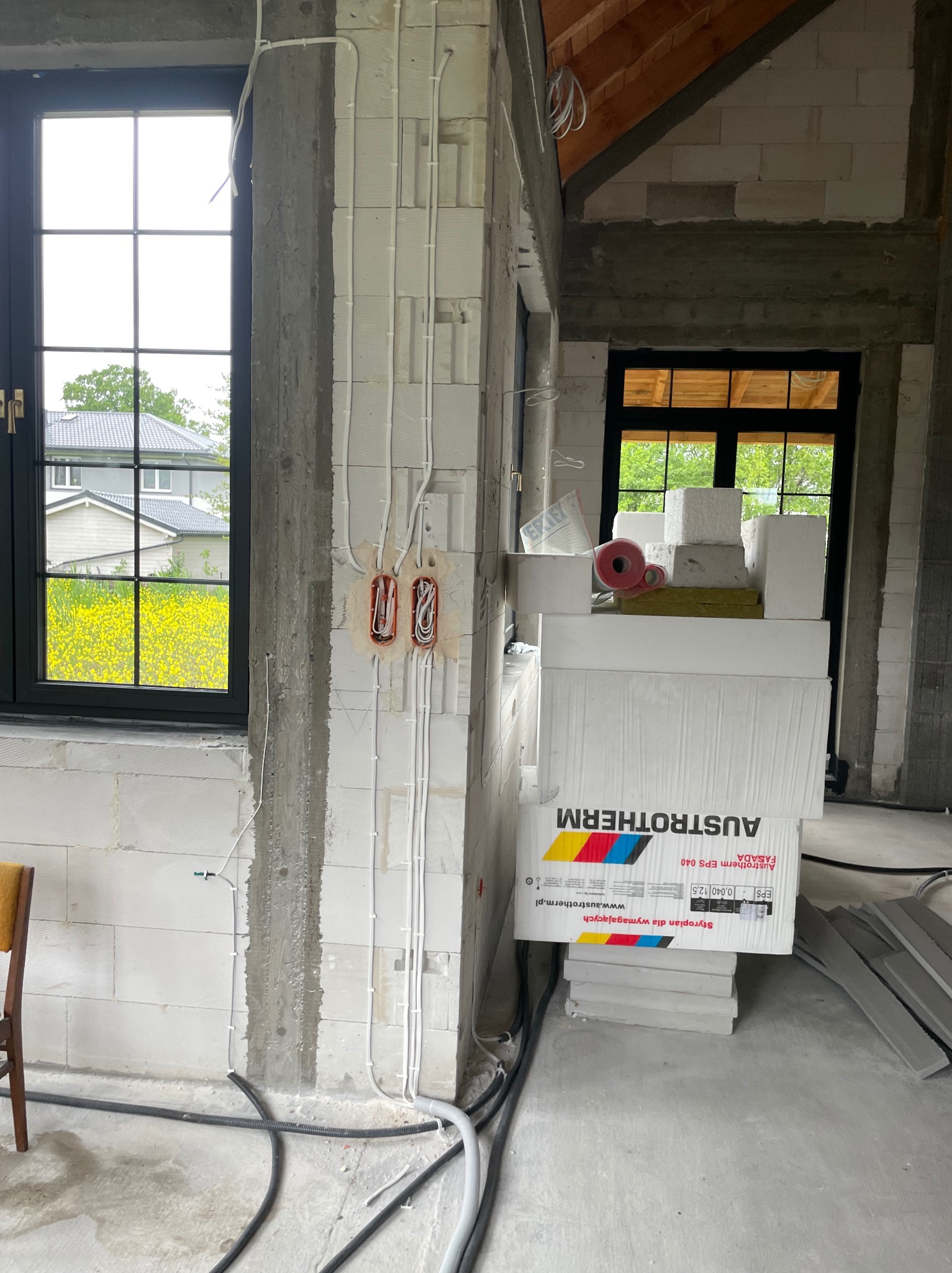 Surowy stan budynku w trakcie prac: widoczne kable elektryczne w ścianie z betonu komórkowego, puszki instalacyjne i okno z widokiem na zieleń.