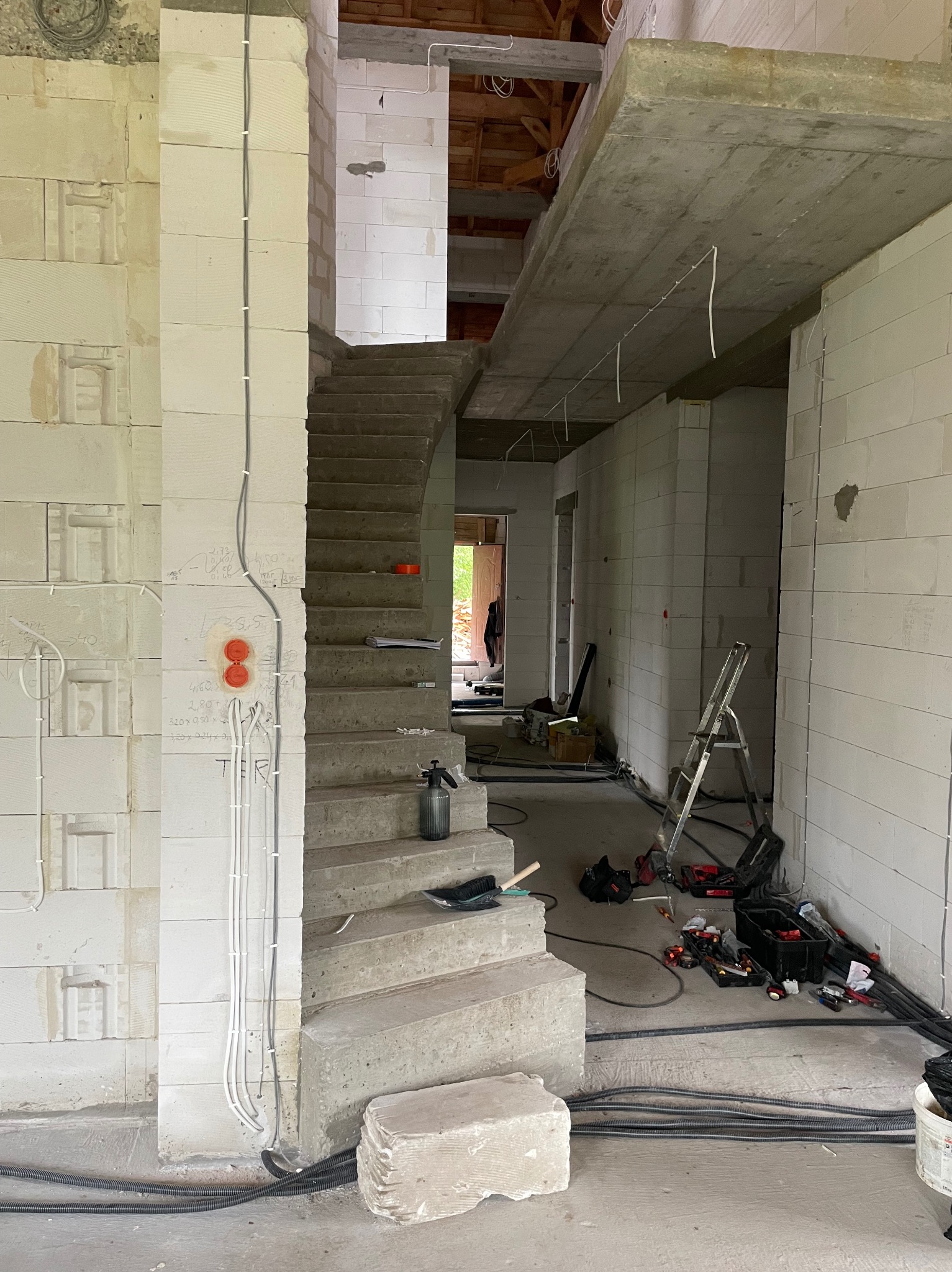 Surowy stan wnętrza w trakcie budowy: ściany z betonu komórkowego, betonowe schody, instalacja elektryczna w peszlach, narzędzia i drabina. Widok na klatkę schodową.
