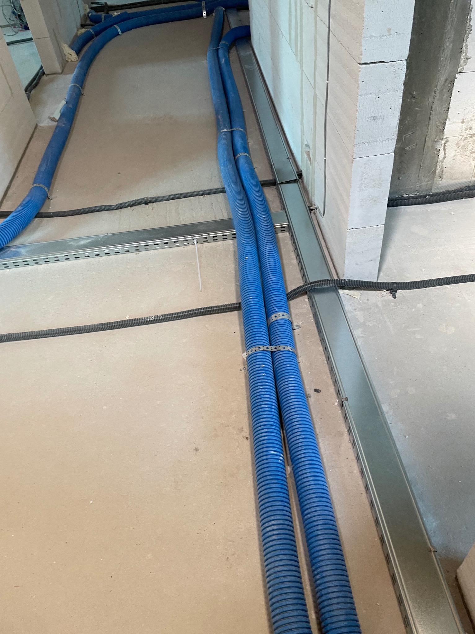 Niebieskie rury w karbowanej osłonie, prowadzone wzdłuż ściany na metalowych wspornikach, czarne kable w peszlach. Instalacja w surowym wnętrzu.