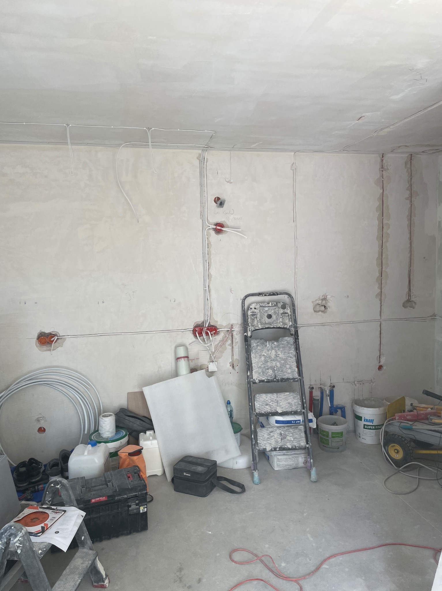 Surowy stan wnętrza w trakcie prac elektrycznych: widoczne kable na ścianach, puszki instalacyjne, drabina i narzędzia. Ściany w szarym tynku, podłoga betonowa.
