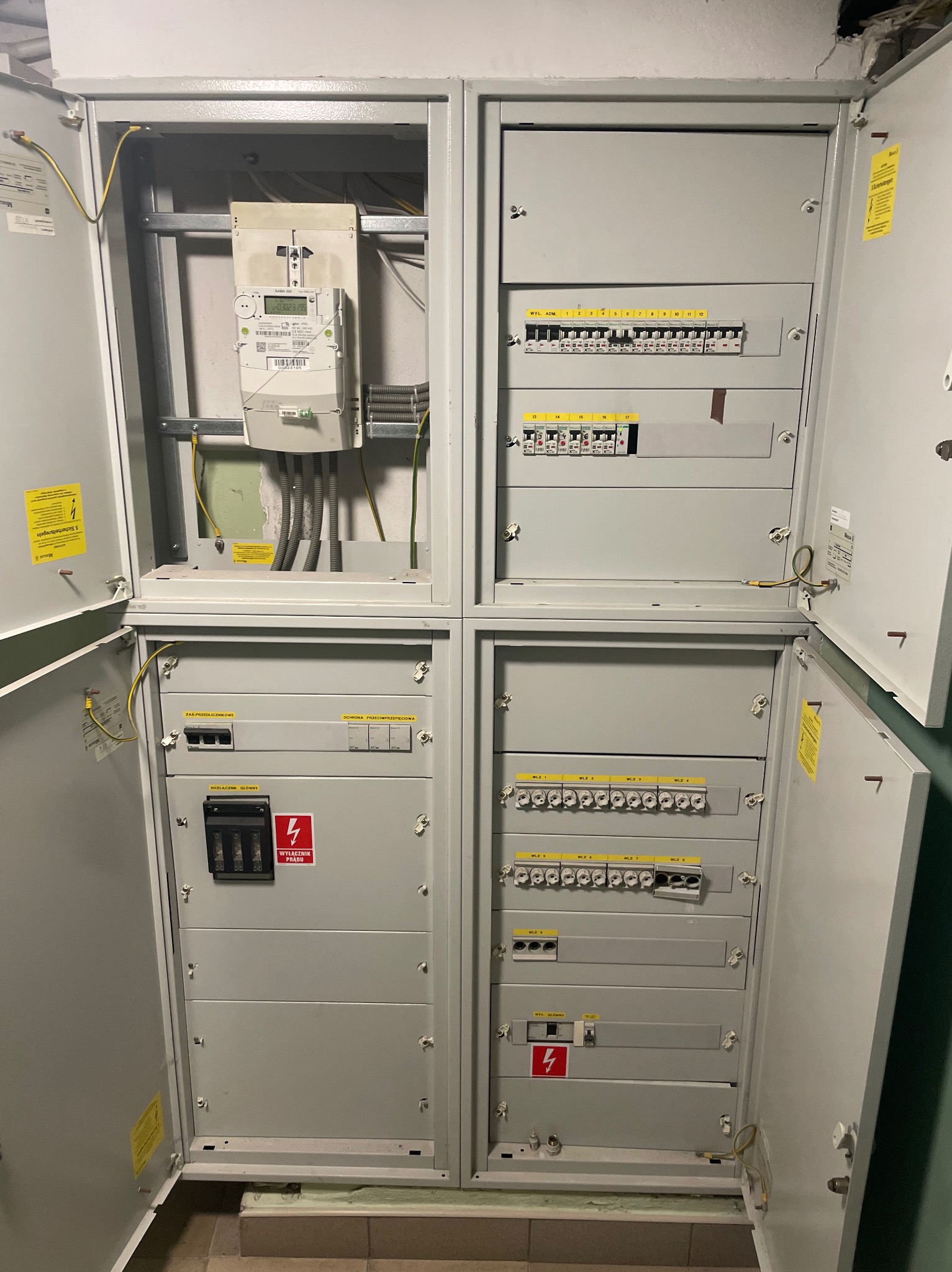 Otwarta, szara szafa elektryczna z licznikiem i bezpiecznikami, widoczne kable i oznaczenia. Instalacja wewnątrz budynku, oświetlenie górne.
