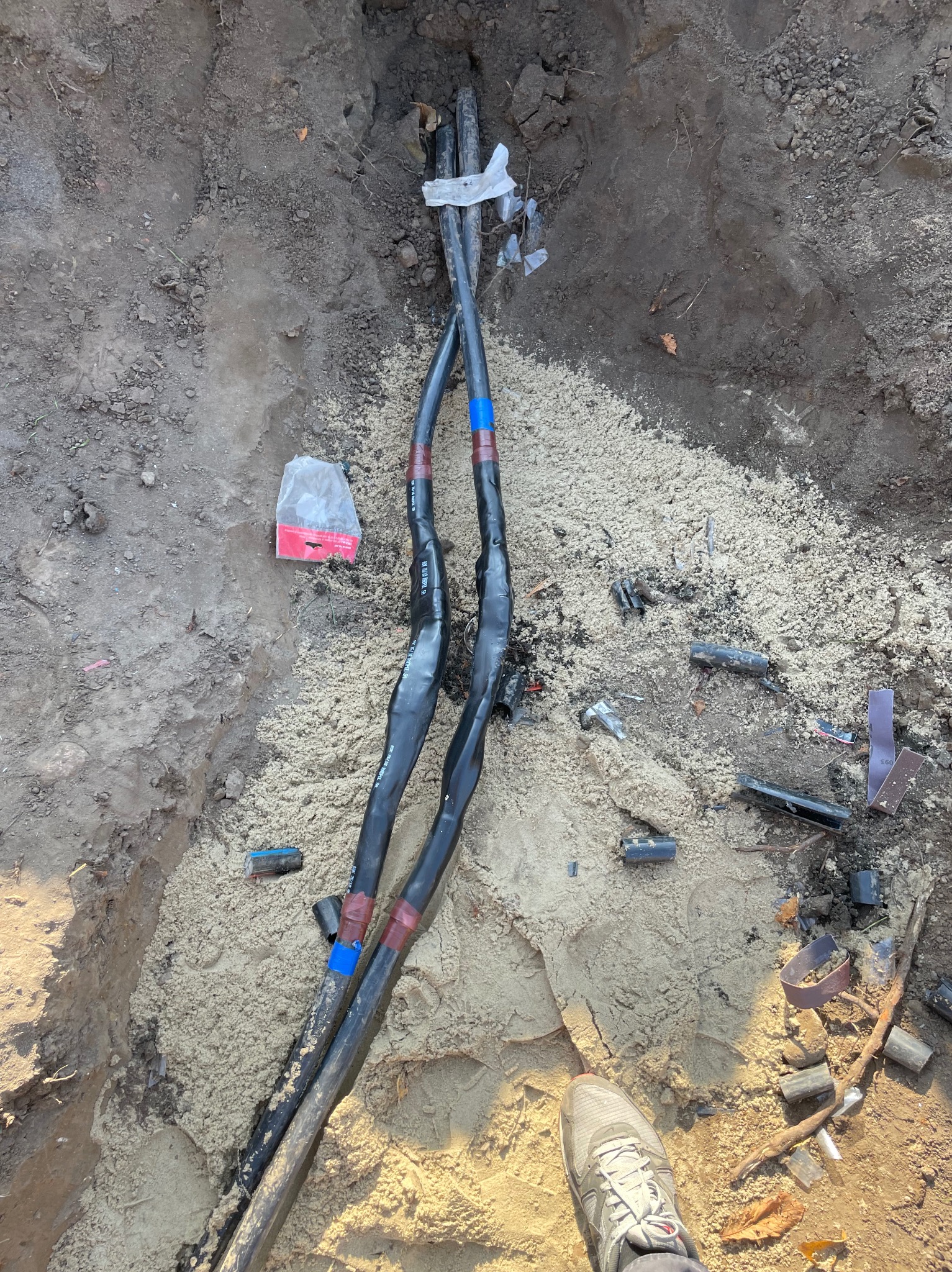Dwa czarne kable elektryczne w wykopie, zabezpieczone taśmą izolacyjną, otoczone piaskiem i elementami montażowymi. Widoczny fragment buta osoby nadzorującej.