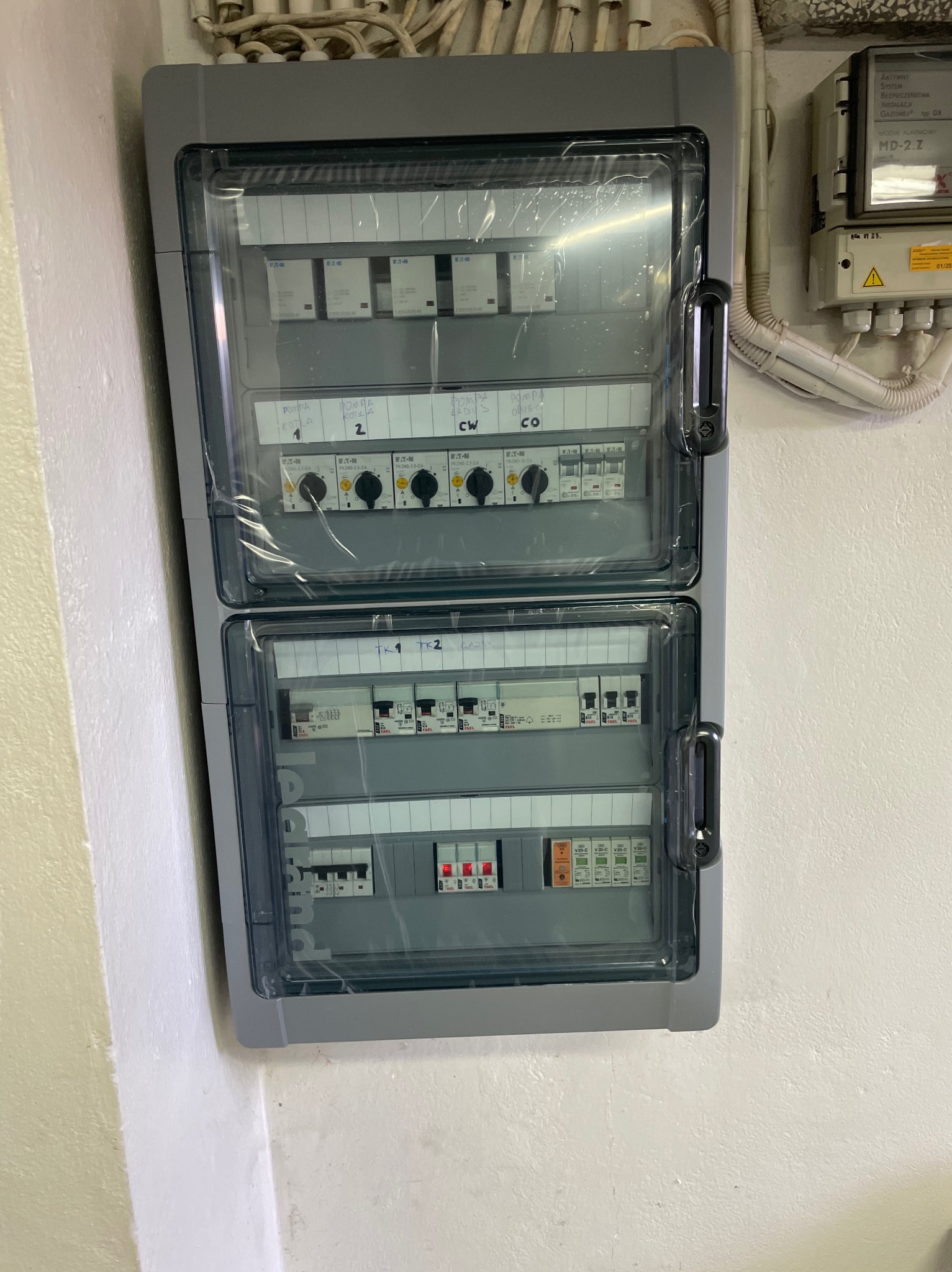 Dwuczęściowa, szara skrzynka elektryczna z przezroczystymi drzwiczkami, zamontowana na białej ścianie. Widoczne bezpieczniki, przełączniki i okablowanie. Obok licznik gazowy.