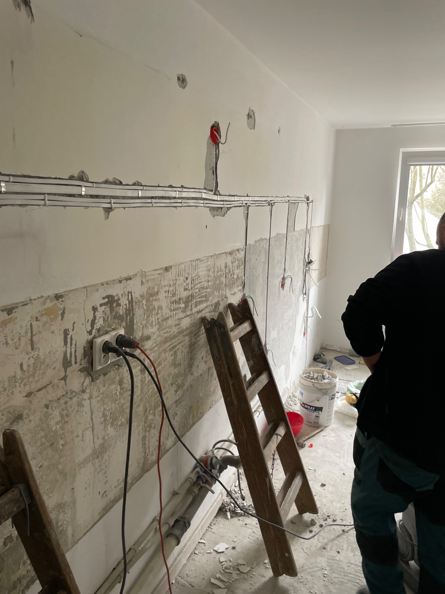 Remont kuchni: widoczne przewody elektryczne zamontowane na ścianie, gniazdko z podłączonymi kablami, drabina i wiadro z narzędziami. Etap prac przed wykończeniem.