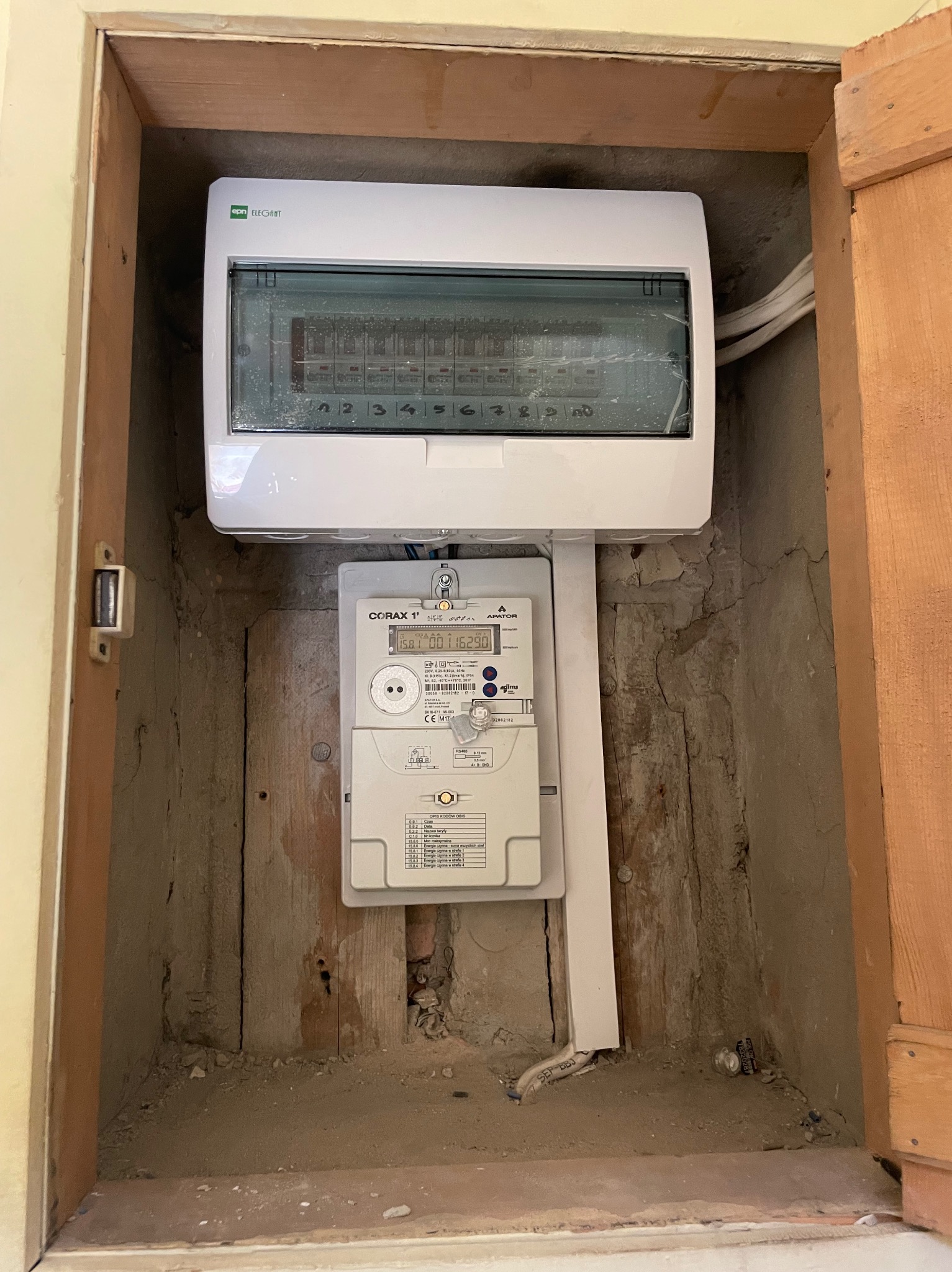 Zamontowana skrzynka elektryczna z licznikiem Corax 1' w drewnianej wnęce. Widoczne kable i elementy instalacji. Ściany wnęki surowe, z widocznymi śladami prac budowlanych.