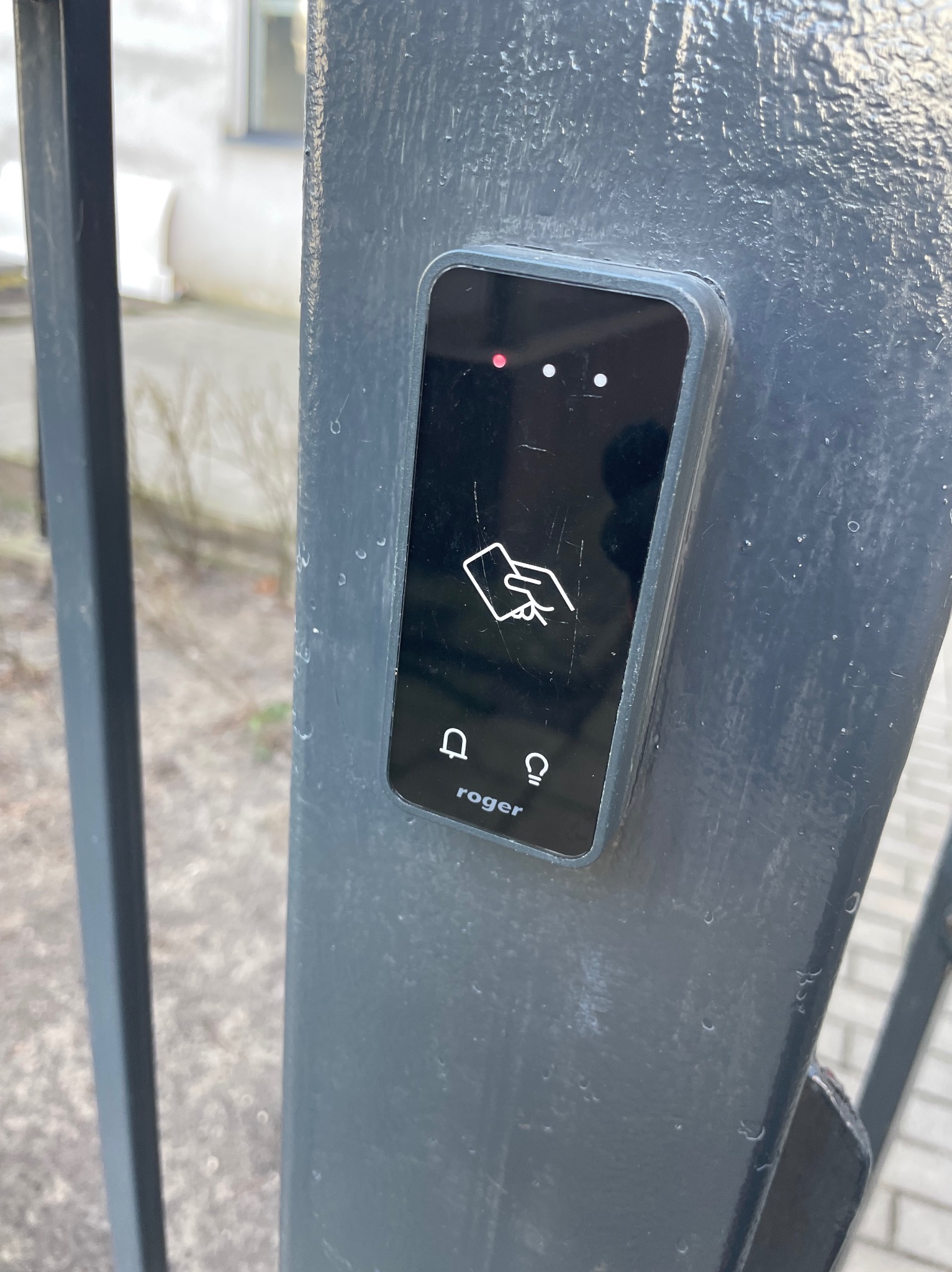 Czytnik kart zbliżeniowych Roger zamontowany na metalowym słupku ogrodzenia, z widocznymi ikonami dzwonka i oświetlenia. Drobne rysy na obudowie.